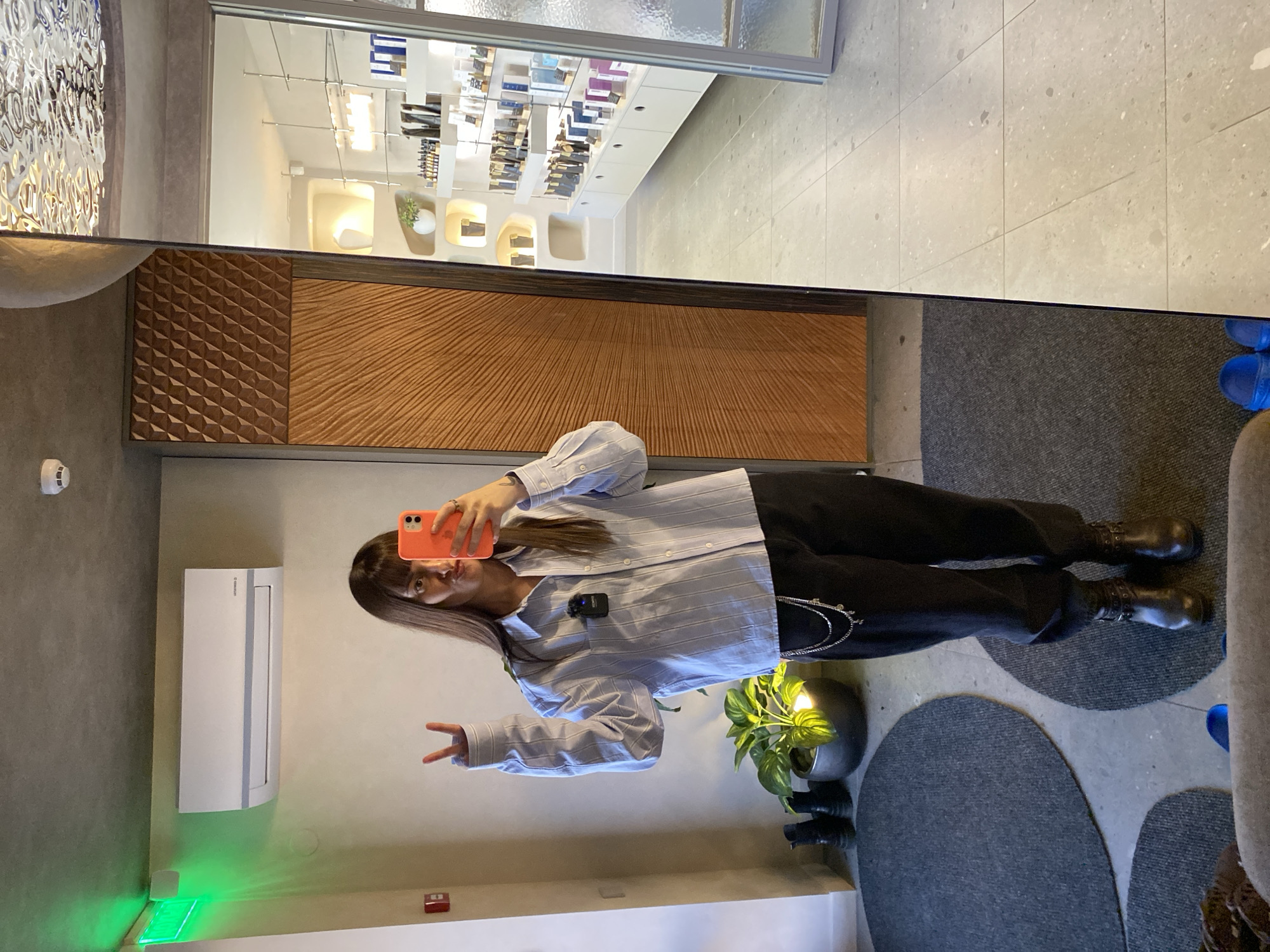 A full-length mirror selfie shows a woman in an indoor setting, likely a retail space. The woman is wearing a light blue, striped button-down shirt, black pants, and dark-colored boots with straps. She is holding an orange smartphone up to her face, using it as a mirror to take the picture. She is giving a peace sign with her left hand.
The environment in the reflection includes a display of retail goods, possibly perfumes, arranged on shelves. The mirrored interior of the store is bright and modern, with geometric wood paneling on the adjacent wall.
Outside the mirror, a neutral-colored wall features an air conditioning unit and various wall fixtures. At the bottom left is a green LED exit sign. Beside the woman is a plant in a gray pot.
The location information, "Izhevsk, Russia," suggests this scene is within a retail space in that city.