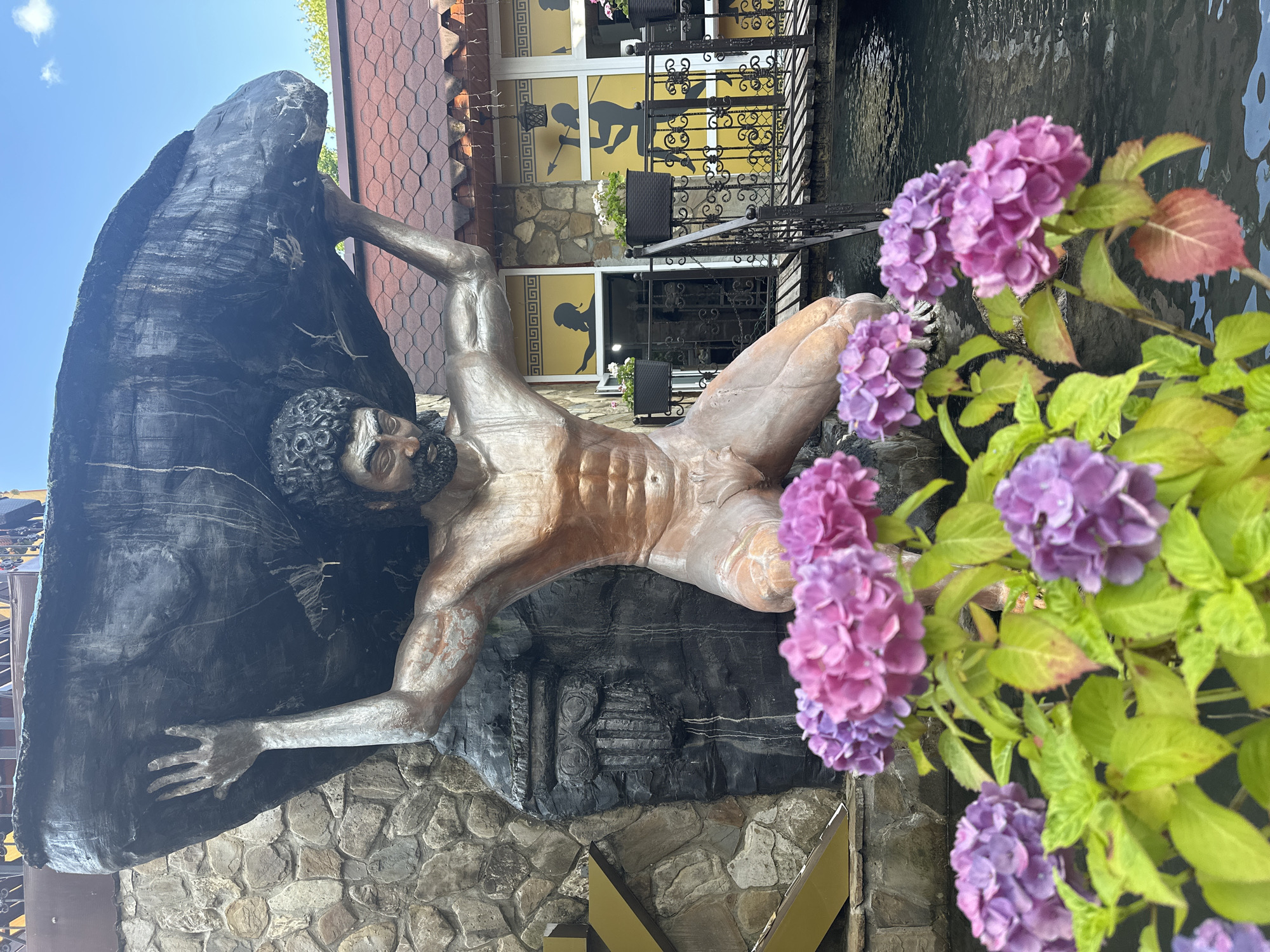 The image shows a statue of Atlas, a Greek mythological figure, in the city of Krasnaya Polyana, Russia. Atlas is depicted kneeling and holding a large rock above his head. The statue is made of bronze and is located in a garden with hydrangeas in the foreground. In the background, there is a building with yellow walls and a tiled roof. The building has windows with silhouettes of people on them. There is also a wooden bridge over a body of water. The sky is blue and there are some clouds. The overall scene is peaceful and picturesque.
