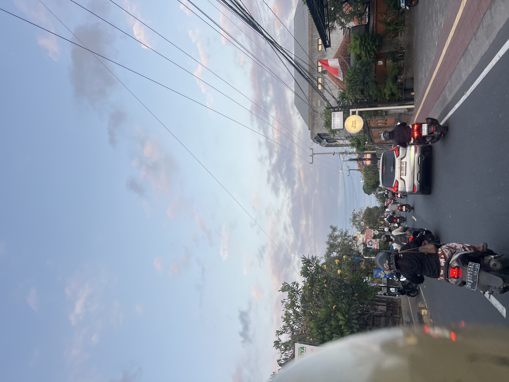 A street scene in Denpasar, Indonesia, captures a moment of evening traffic. The sky is a pale blue with scattered, wispy clouds tinged with pink and gray, suggesting either sunrise or sunset. Overhead, a dense network of dark electrical wires crisscrosses the frame, adding a sense of urban clutter.
The perspective is from someone on a motorcycle, looking forward. In the foreground, a portion of a light-colored motorcycle helmet is visible on the left. The road is asphalt, marked with white and yellow lines. A steady stream of vehicles, predominantly motorcycles, moves along the street. Several motorcycles are in motion, with riders wearing helmets and casual clothing. One motorcycle in the mid-ground has a rider wearing a black hoodie and a patterned skirt. Another motorcycle to the right has a rider in a black jacket.
A silver SUV is also present in the traffic, its license plate reading "DK 1595 QP". On the right side of the road, a sidewalk runs alongside buildings. One building prominently displays a red and white Indonesian flag. Above the sidewalk, signs are visible, including one that reads "DOKTER GIGI DENTIST" and another with "Rasa Beauty". Lush green trees with yellow flowers line the street on the left and partially obscure some of the buildings on the right. The overall atmosphere is that of a typical, active urban street during a busy time of day, illuminated by the soft, diffused light of dusk.
