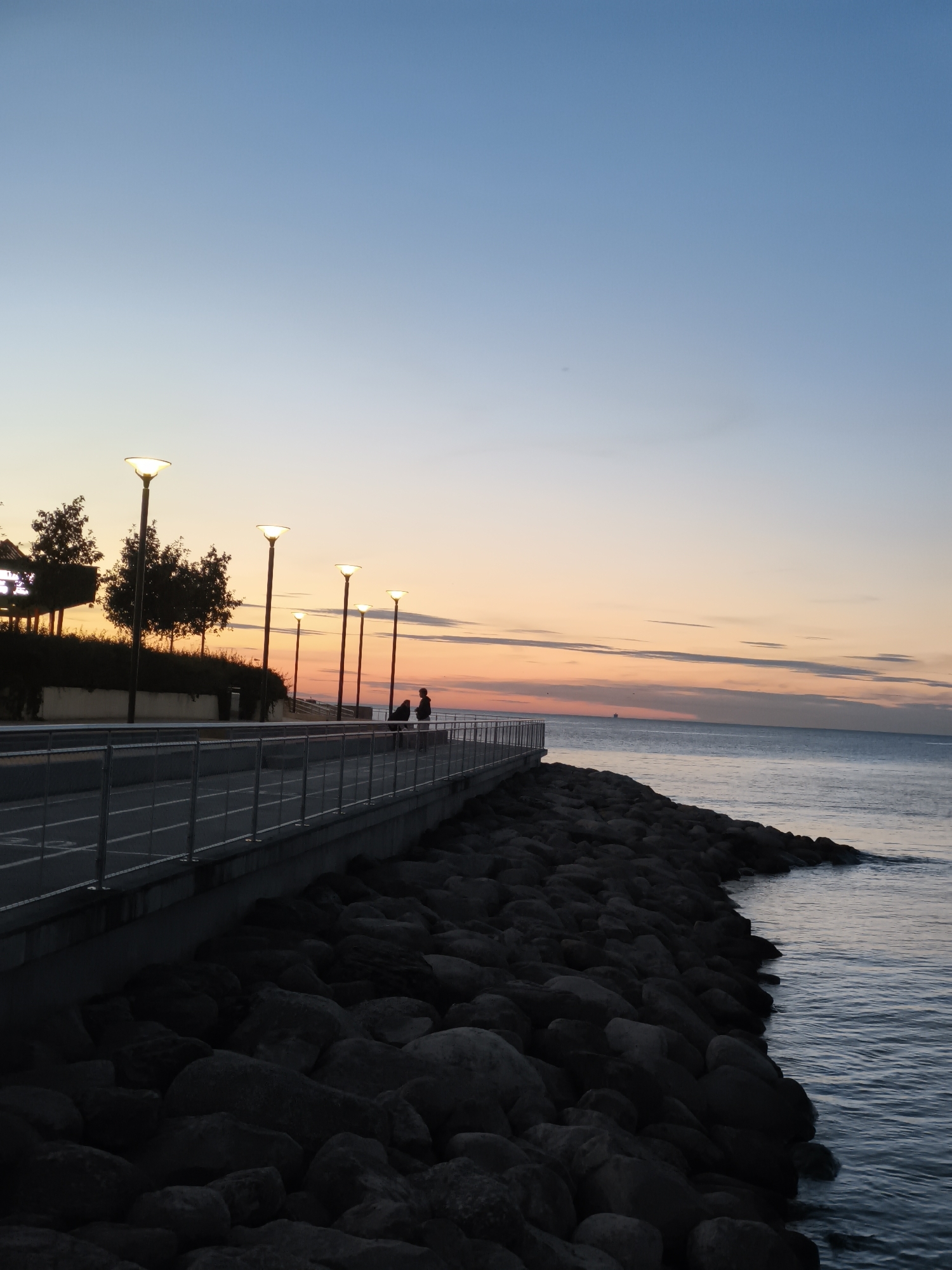 The image depicts a sunset scene along the coast of Tallinn, Estonia. A paved promenade with a metal railing runs parallel to a rocky shoreline. Several lampposts illuminate the promenade. Two individuals are silhouetted on the promenade, facing the water. The sky transitions from a pale blue at the top to hues of orange and pink near the horizon, where a calm sea stretches out. A distant vessel is visible on the water. The rocky area in the foreground is dark and densely packed with stones of various sizes, with the sea gently lapping at their edges.