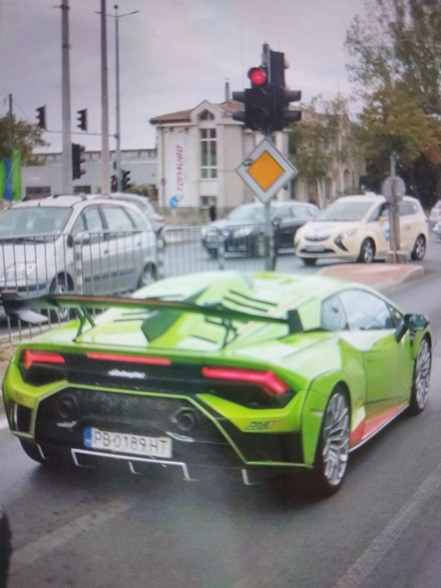 The image shows a green Lamborghini Huracán with license plate "PB-0189 HT" on a city street. A traffic light shows red. Cars are behind the Lamborghini. A building with a large banner that reads "FORMURO" is in the background. The location is Plovdiv, Bulgaria.