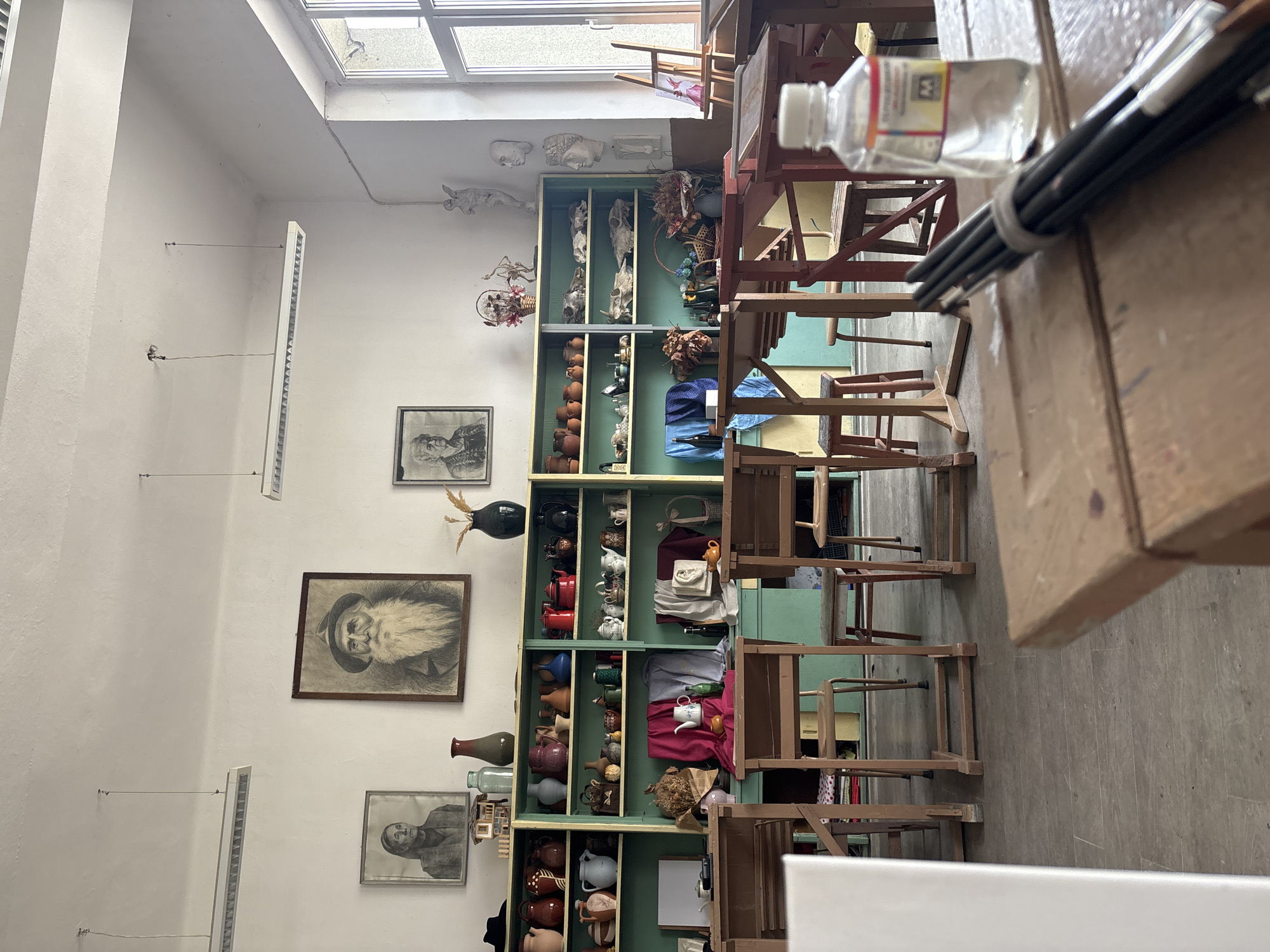 This image shows the interior of an art studio or classroom. The walls are white and adorned with artwork, including framed portrait sketches. A tall, green display shelf is stocked with various objects like pottery, decorative items, and what appear to be casts or sculptures. There are several wooden chairs, some stacked, suggesting a workspace. A collection of paintbrushes and a container are visible in the foreground. It appears to be a studio in Troyan, Bulgaria.
