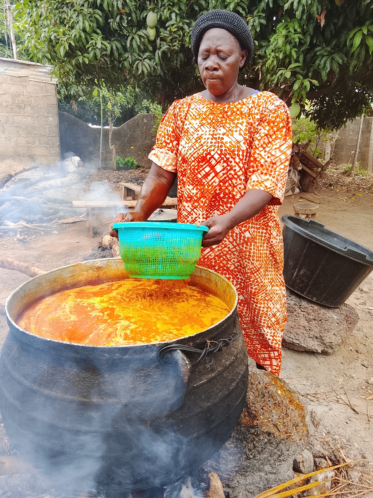 In Akwanga, Nigeria, an older woman wearing a patterned orange dress and a black head covering stands next to a large, soot-covered pot filled with a yellow-orange liquid. She is holding a green plastic colander over the pot, with liquid dripping from it into the pot. Smoke rises from underneath the pot, which is supported by rocks on the ground. In the background, there is a concrete structure, a tree with green leaves, and a black plastic tub. The ground is mostly bare dirt.