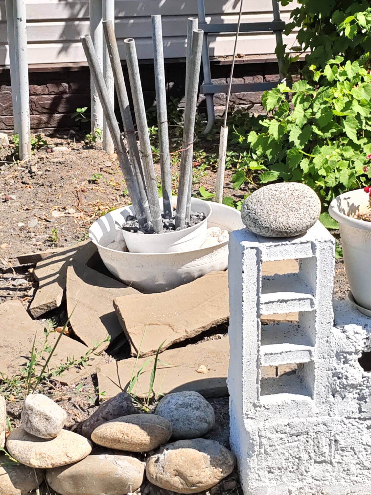 This is a close-up, outdoor, eye-level shot of a small garden scene. In the foreground, several rounded, smooth stones of varying sizes are clustered on the ground. To the right, a white, rough-textured cinder block is standing upright, topped with a single, rounded grey stone. Behind the cinder block, a white planter with a similar grey stone on top is visible. To the left, a large white planter holds several thin, grey metal poles, some of which are tied together with red string. The planter is sitting on a foundation of large, flat, light brown stones. In the background, a section of a building with horizontal siding and a brick-like lower portion is visible. Lush green foliage with large leaves is growing on the right side of the frame, partially obscuring the building. The ground is covered in dirt and small stones. The lighting suggests a sunny day, with distinct shadows cast by the objects. The scene is likely a residential garden in Rostov-on-Don, Russia, given the context provided.