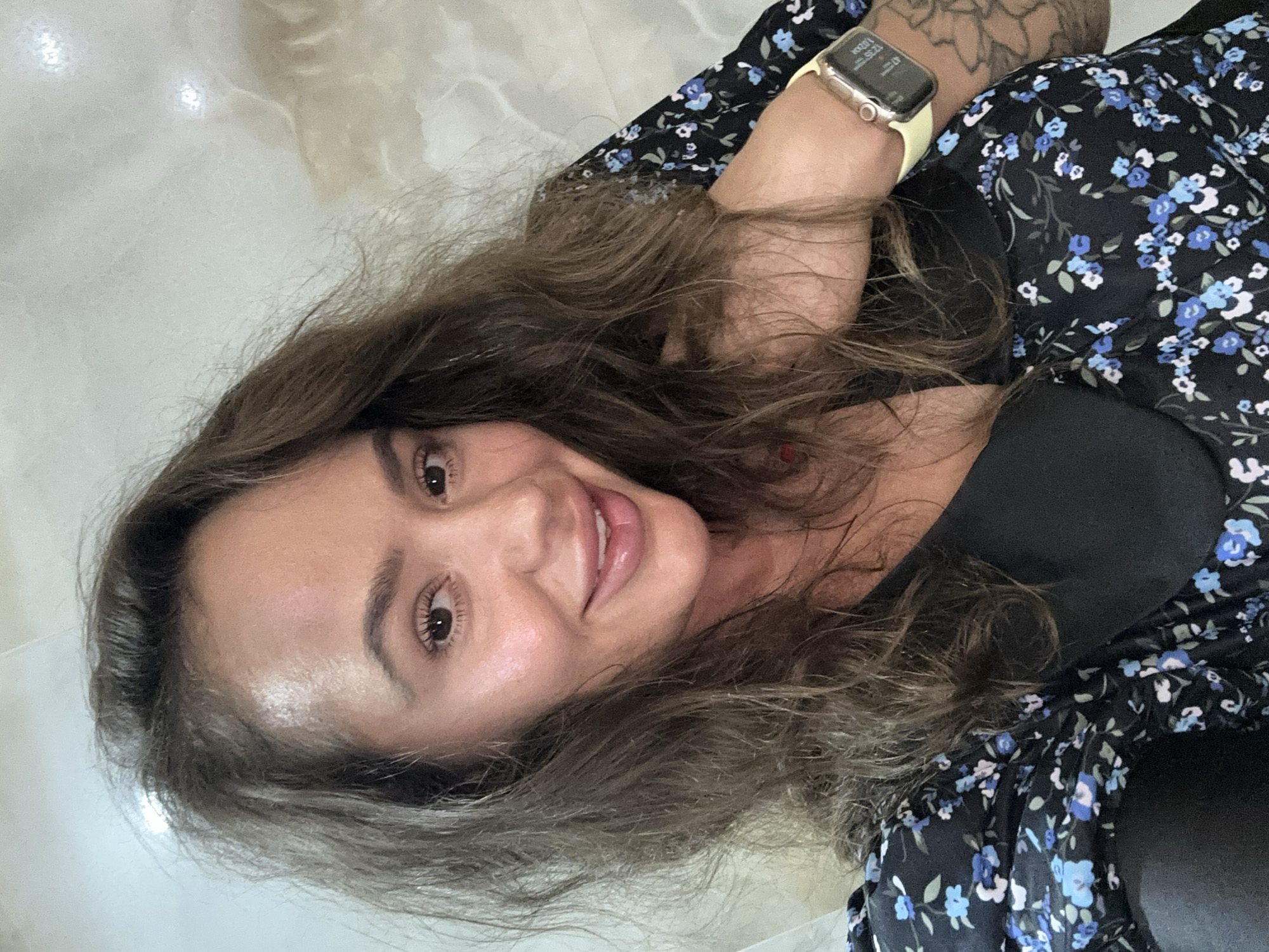 A woman is posing for a selfie indoors, likely in a home environment. She is wearing a black dress with a blue floral pattern. A smartwatch is visible on her left wrist. The background appears to be a tiled wall with a marble-like pattern. The woman has medium-length brown hair, brown eyes, and is smiling. There is no indication of the location being in Duvankul', Russia.