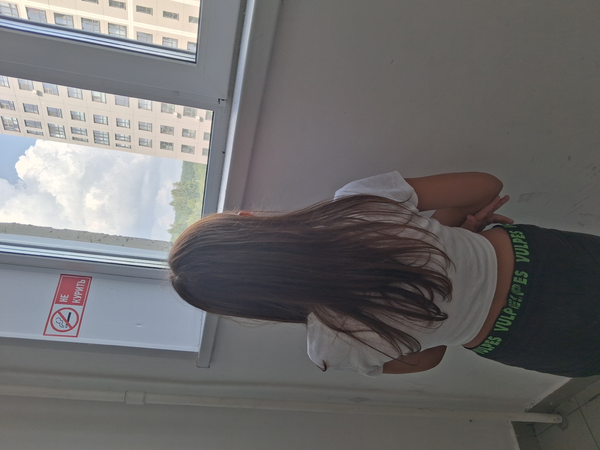 The image shows a young person, viewed from behind, standing by a window and looking out. The person has long, dark brown hair and is wearing a white cropped t-shirt and dark shorts with green lettering that appears to say "VULPES" repeated. To the left of the person, on the window frame, is a red sign with a white border depicting a cigarette with a line through it, and text in Russian that reads "НЕ КУРИТЬ" (DO NOT SMOKE). Through the window, a tall, modern apartment building with many windows is visible, along with a cloudy sky and some trees in the distance. The surrounding environment appears to be an indoor hallway or stairwell, with a light-colored wall and a floor with visible grout lines. The scene is consistent with an interior view from a residential building in Novosibirsk, Russia.