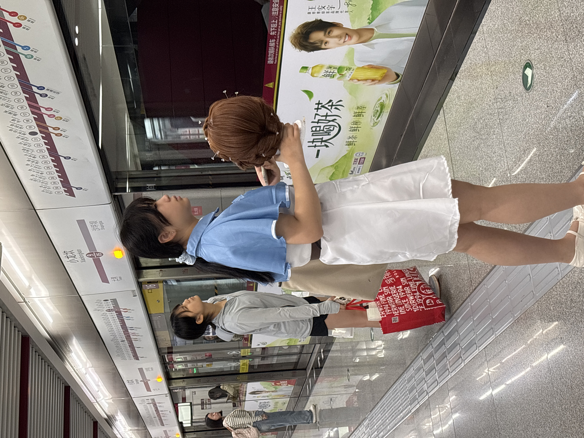 Here is a forensic description of the image.
The image shows the inside of a subway station. In the foreground, a person is standing on the platform with a mannequin head. The mannequin head is brown with pins in the hair. The person is wearing a blue top and a white skirt. Behind the person, there is another person standing on the platform. This person is wearing a gray sweatshirt and black shorts, and holding a red tote bag. There are advertisements posted on the wall of the subway station, including an ad for a beverage. A subway map with station names and numbers is visible.