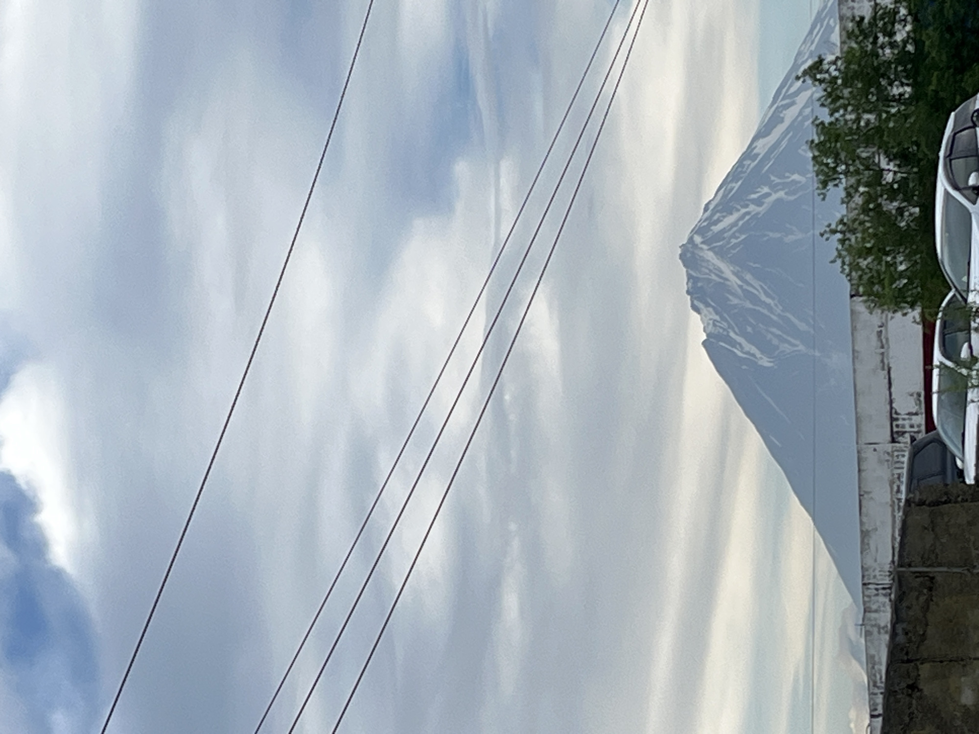 The image captures a distant view of a snow-capped volcano, likely one of the volcanoes visible from Petropavlovsk-Kamchatsky, Russia, such as Avachinsky or Koryaksky. The volcano dominates the background, with patches of snow streaking down its slopes against a muted blue and white cloudy sky. In the foreground, a low, weathered concrete structure runs horizontally across the frame. Behind this structure, green trees are visible. Several power lines crisscross the sky in the upper portion of the image. The lower part of the image shows parts of at least two cars parked behind the concrete barrier. The overall scene suggests an overcast day.