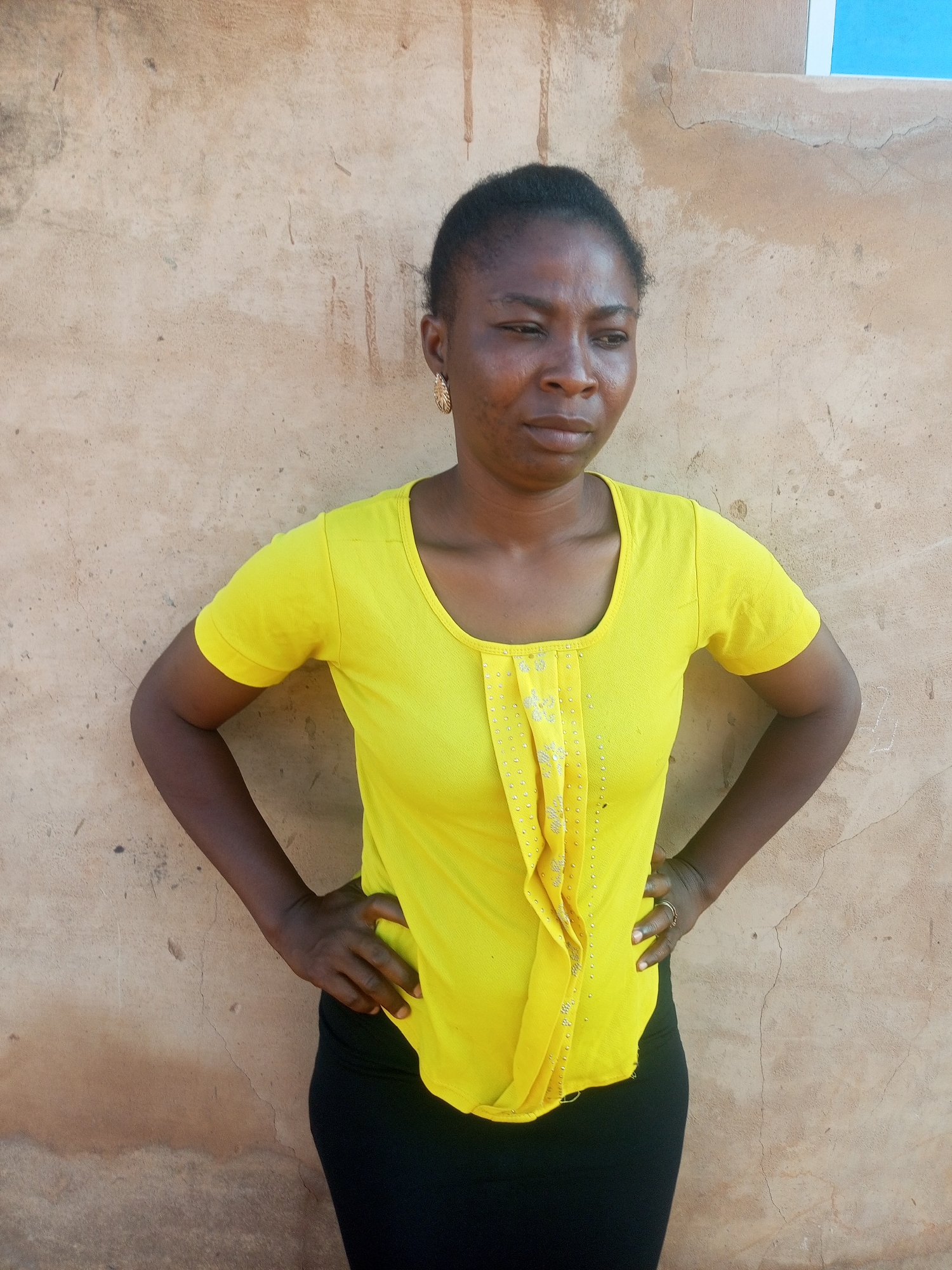 A full-body shot captures a Black woman standing outdoors in front of a textured, light brown wall. She has dark hair pulled back and is wearing a bright yellow, short-sleeved t-shirt with decorative pleats and embellishments on the chest, and a black skirt. Her hands are placed on her hips. A small part of a light blue object is visible in the upper right corner of the frame. The lighting suggests it is daytime, with shadows indicating natural light. The wall shows signs of wear, including cracks and some staining. The environment appears to be a simple outdoor setting, possibly a residential area in Tempure, Nigeria, given the architectural style of the wall.