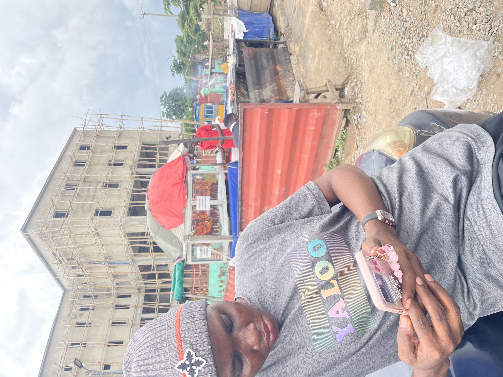 The image shows an outdoor scene in Galadimawa Village, Nigeria, with a partially constructed building in the background. In the foreground, a person wearing a grey beanie and a grey t-shirt with "YALOO" printed on it in colorful letters is looking at a smartphone in their hands. The smartphone has a pink case decorated with charms and beads. The person is sitting on what appears to be the hood of a vehicle. Behind the person, there are several food stalls. One stall has a red umbrella and displays grilled chicken. Another stall features a person in a red shirt standing near a grill with food cooking. The ground is a mix of gravel and dirt, with some vegetation visible. A crumpled plastic bag lies on the ground to the right. The sky is overcast.