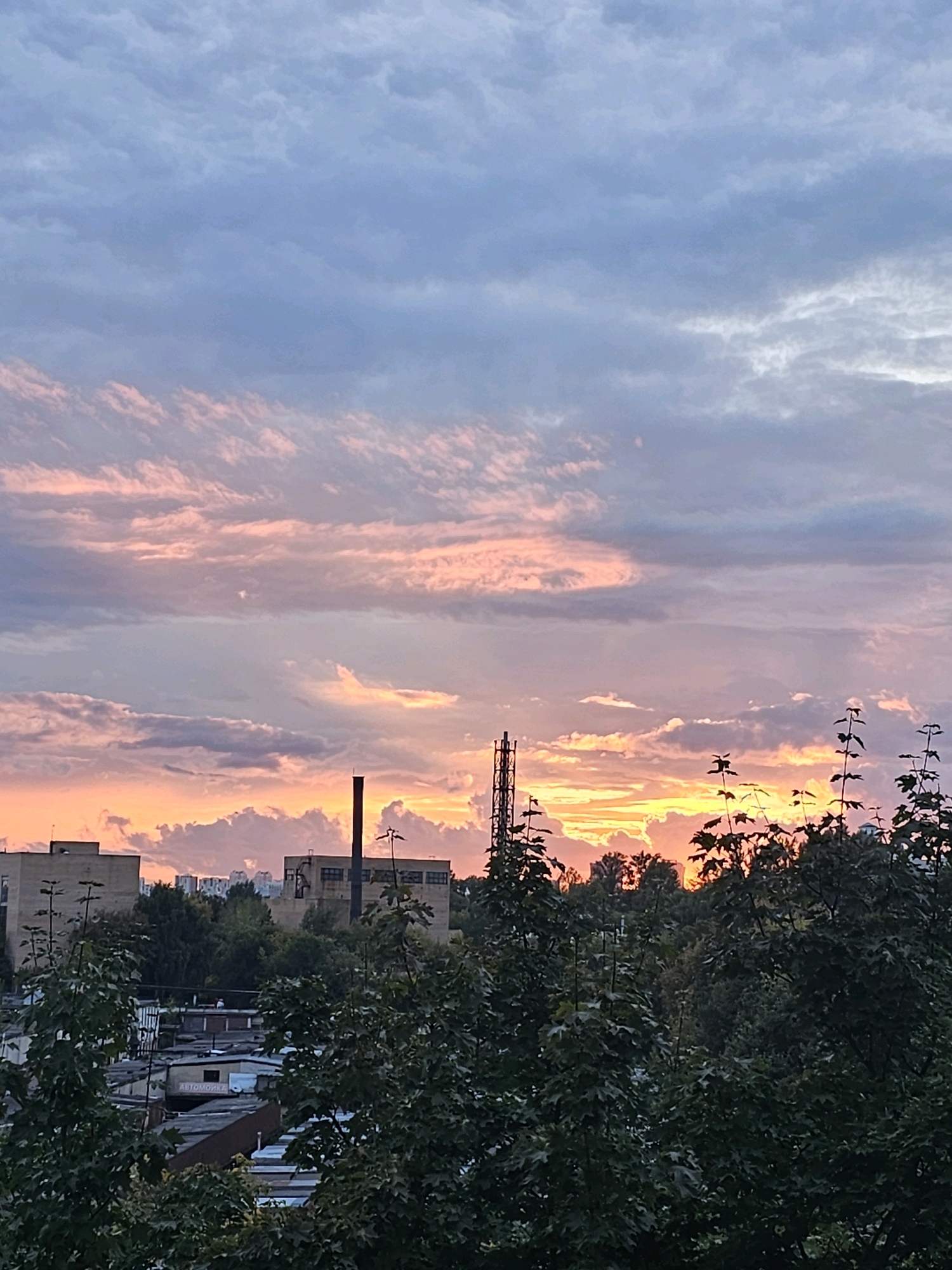 The image depicts a cityscape at dusk in Khimki, Russia. A vibrant sunset paints the sky with hues of pink, orange, and purple, illuminating scattered clouds. In the foreground, dark green foliage of trees partially obscures the view of industrial buildings and garages below. A tall, slender industrial chimney rises from one of the buildings, silhouetted against the colorful sky. The overall scene conveys a sense of an urban environment transitioning from day to night.