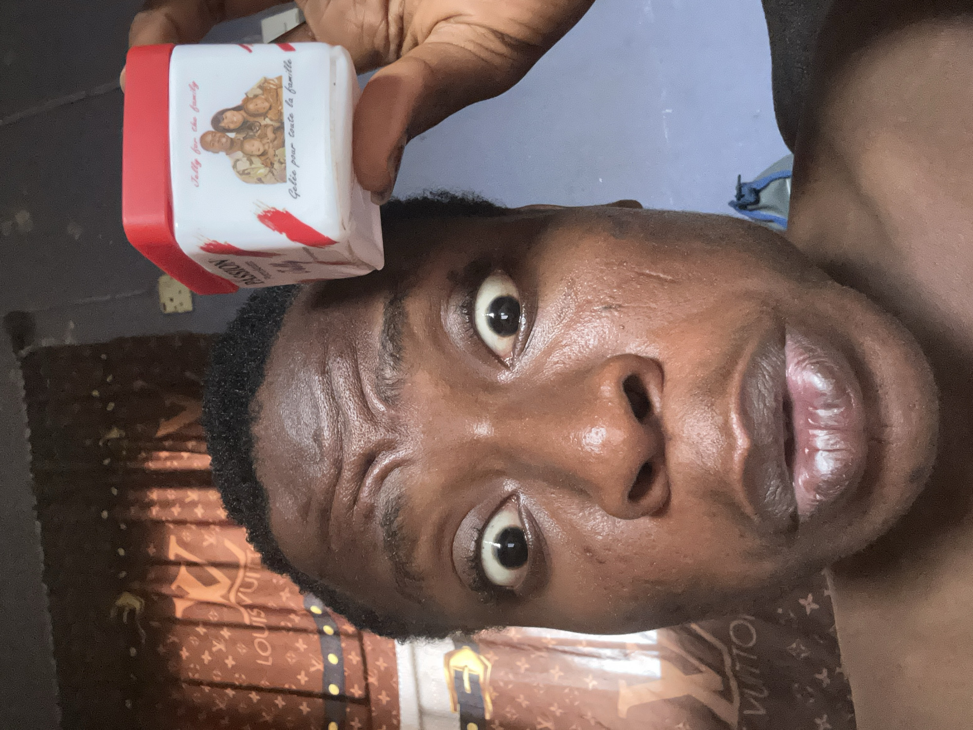 A man is holding a white and red container with the word "Passion" on it near his forehead. The background includes a brown Louis Vuitton-patterned fabric and a blue-grey wall.