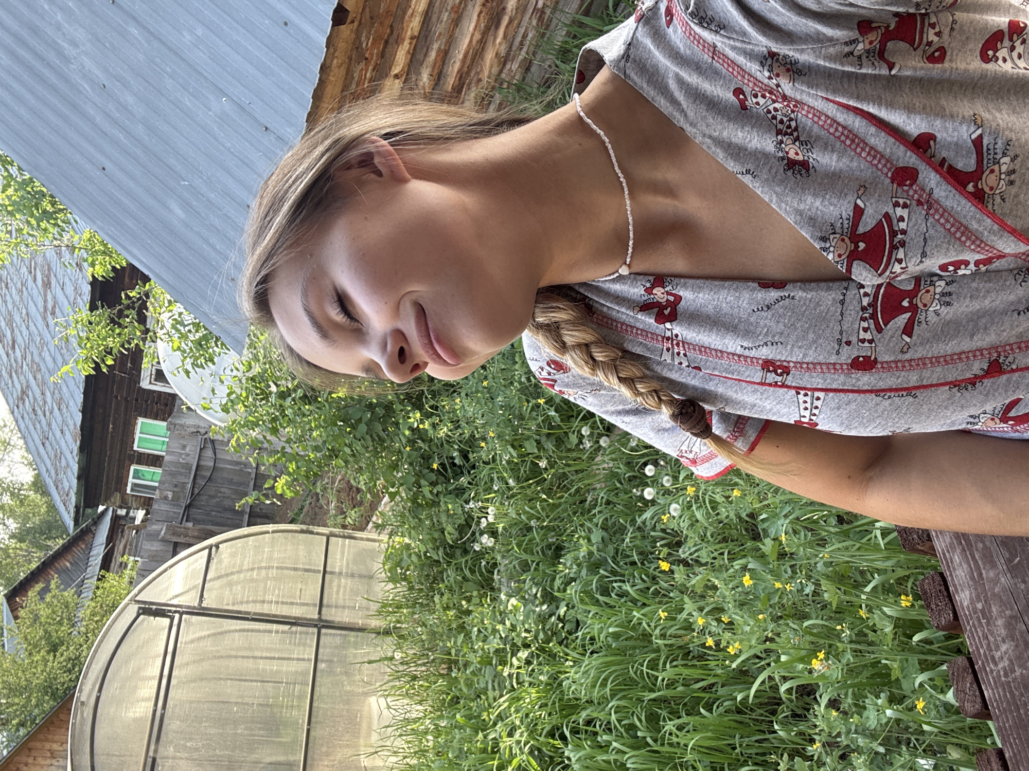 A young woman is the primary subject of the image, her eyes are closed, her blonde hair is braided, and she wears a necklace and patterned grey top. She is in a rural setting, evident from the foliage and a greenhouse-like structure nearby. A wooden building stands in the background, with a shed roofed with corrugated metal on the left. Yellow flowers and dandelions can be seen in the grassy area. The image is taken in a garden, possibly near Chaikovsky, Russia.