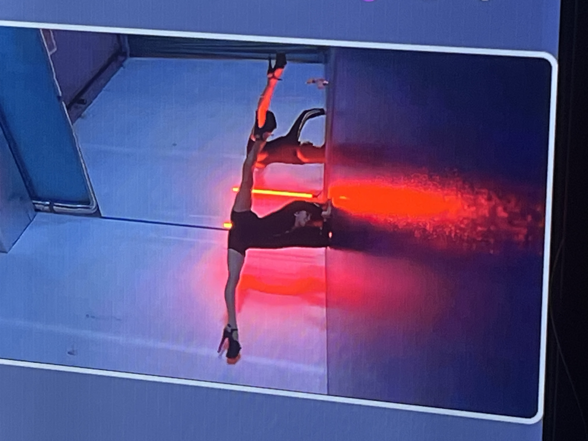 A person is performing a contortionist split pose on a stage with red lighting. The person is wearing black attire and high heels. The background is a blue wall with some structural elements visible at the top. The floor is dark and reflective, showing the red light. The image is a still from a video feed, as indicated by the white border and the framing. The location within the city of Zoriane, Ukraine, is not provided by the visual information in the image.