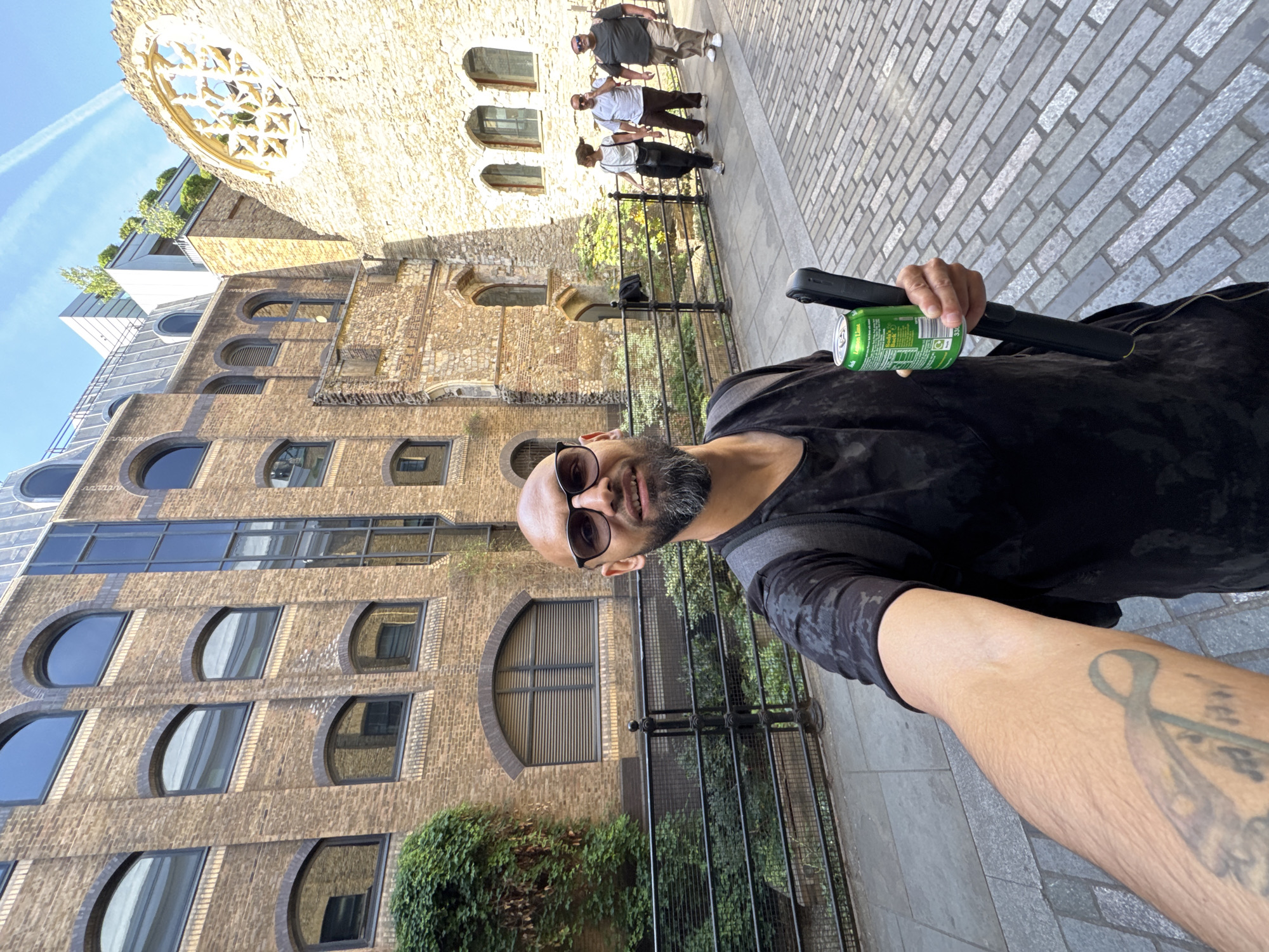 The image is a selfie of a bald man with a beard and sunglasses holding a green soda can and a black selfie stick. He is standing on a paved area, likely a sidewalk. Behind him is an old brick building with arched windows and a round, decorative window at the top. In the background, a few people are walking on the sidewalk near the old building. The modern brick building to the left has semi-circular windows. The location appears to be in London, United Kingdom. The buildings suggest an area with a mixture of historical and modern architecture.