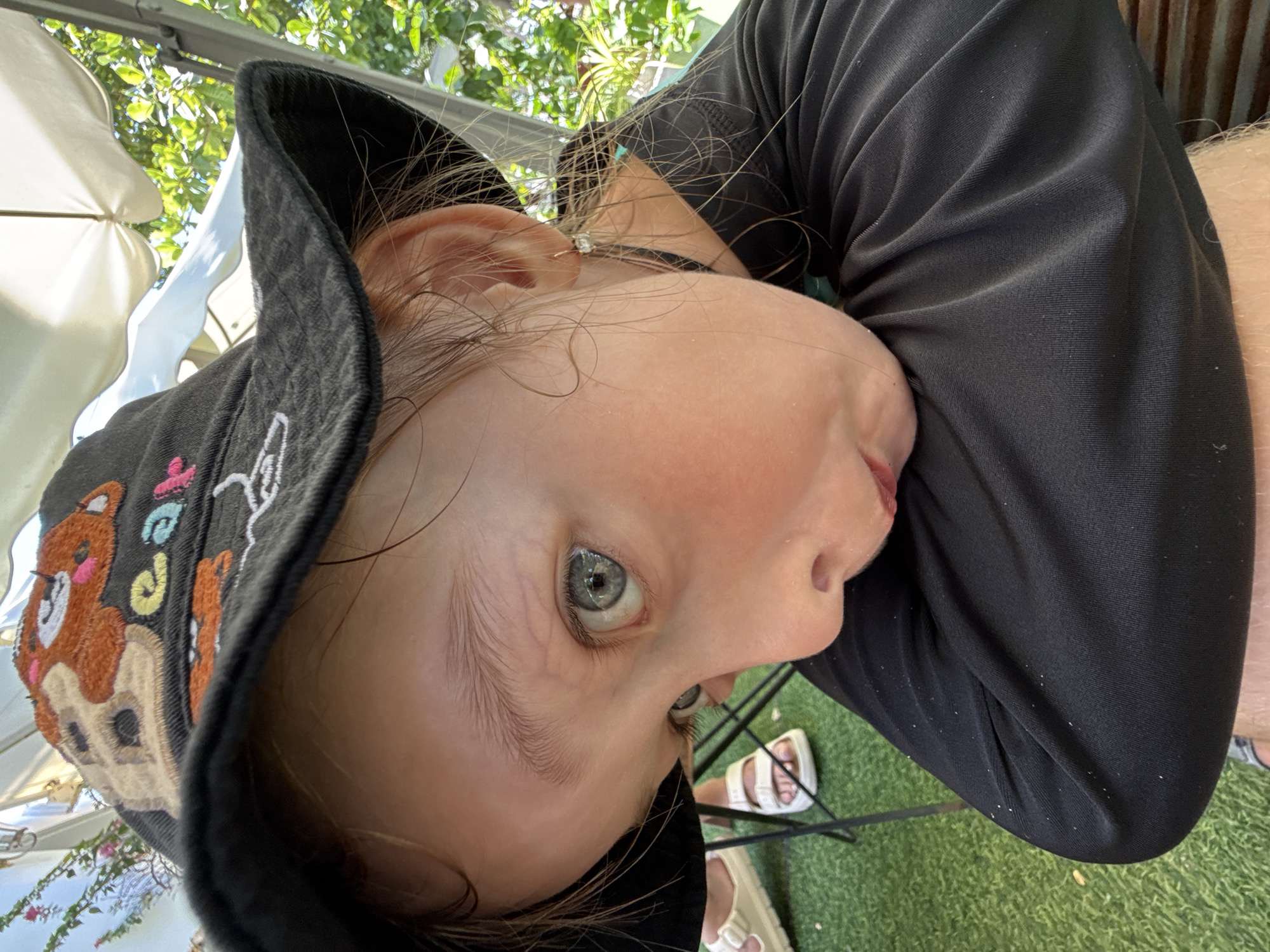 The media file is a close-up, low-angle shot of a young child wearing a black bucket hat with embroidered teddy bear and flower designs. The child's head is tilted, and they are looking upwards. Their left eye is prominent, showing a light blue iris. They are wearing a black long-sleeved shirt, and their arm is visible. The background suggests an outdoor setting, with greenery and a white umbrella visible, implying it might be daytime and sunny. There is no visible text in the image. The child's expression is neutral, and the overall tone is candid. The location context provided is Nha Trang, Vietnam, which is consistent with an outdoor, potentially tropical environment.