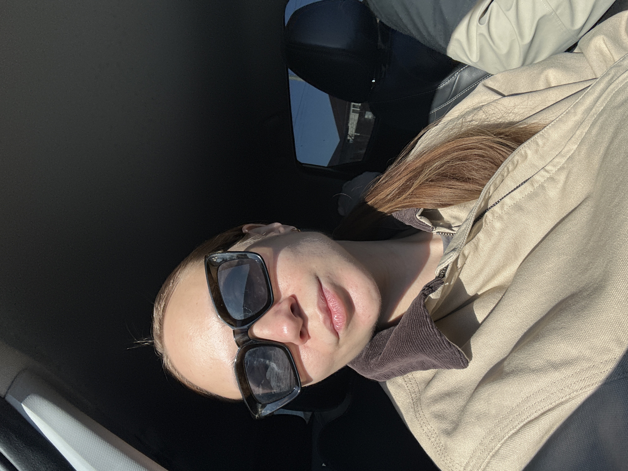 A person with light skin and reddish-brown hair, wearing sunglasses, is seated in a vehicle. The individual is facing the camera at an angle, wearing a light beige jacket over a brown garment. The vehicle's interior is predominantly dark, with black leather seating. A portion of the car window is visible, showing a glimpse of the outside environment with a building and sky. The image does not contain information regarding the location in the city of Kharnavy, Russia.