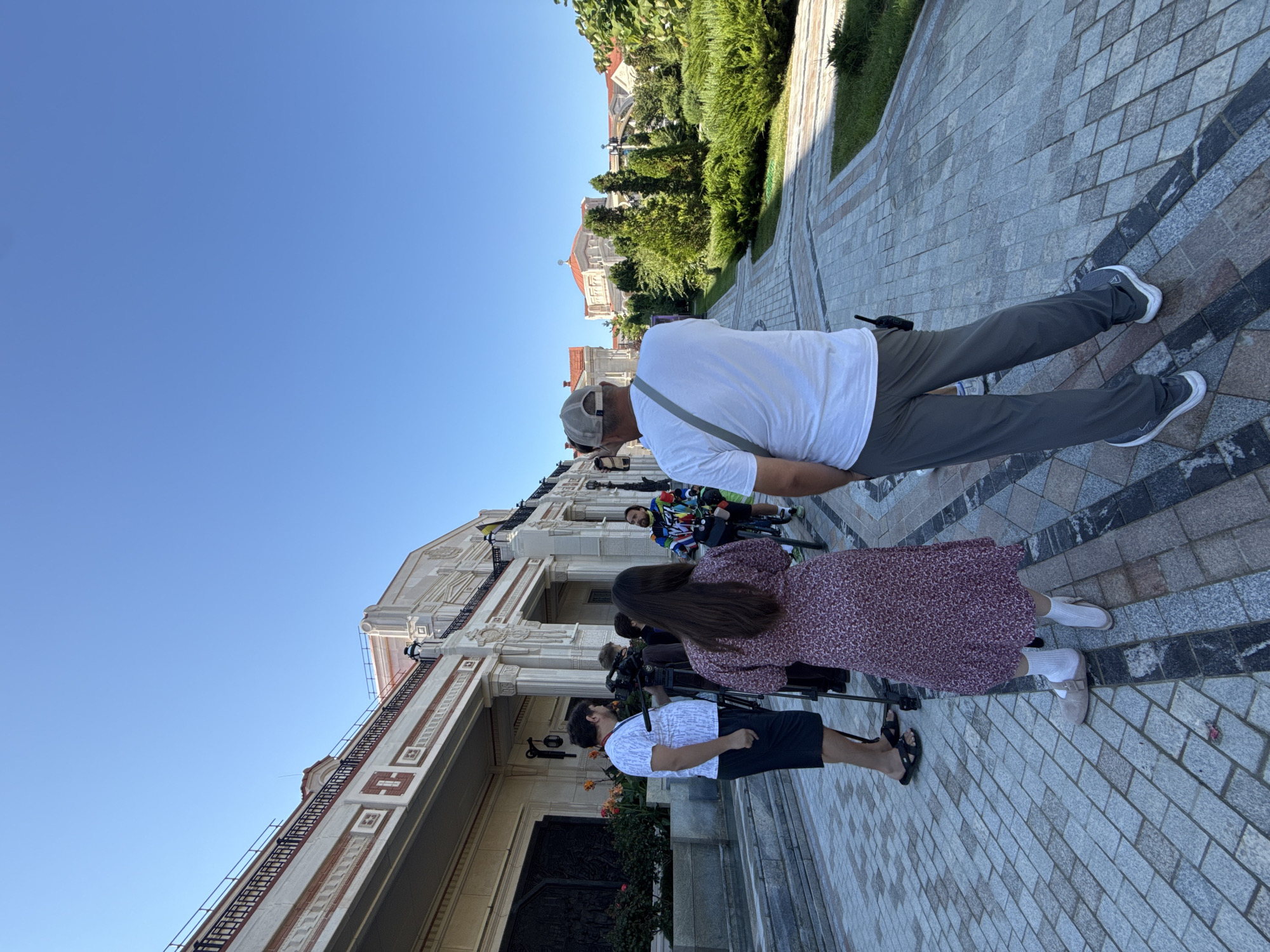 In Sevastopol, Ukraine, outside a grand building with ornate white columns and a red-tiled roof, a group of people are gathered on a patterned paved walkway under a clear blue sky. Several individuals are facing a camera on a tripod, suggesting a media event or filming. One person, wearing a white t-shirt and dark shorts, stands near the building. Another person, a woman in a long, patterned dress, faces away from the camera. A man wearing a white t-shirt and a baseball cap is also in the frame, as is a person on a bicycle wearing a brightly colored jacket. The building appears to be a significant structure, possibly a government building or a cultural institution, with decorative elements and lush greenery surrounding it. The presence of professional camera equipment indicates a formal or public event is taking place.