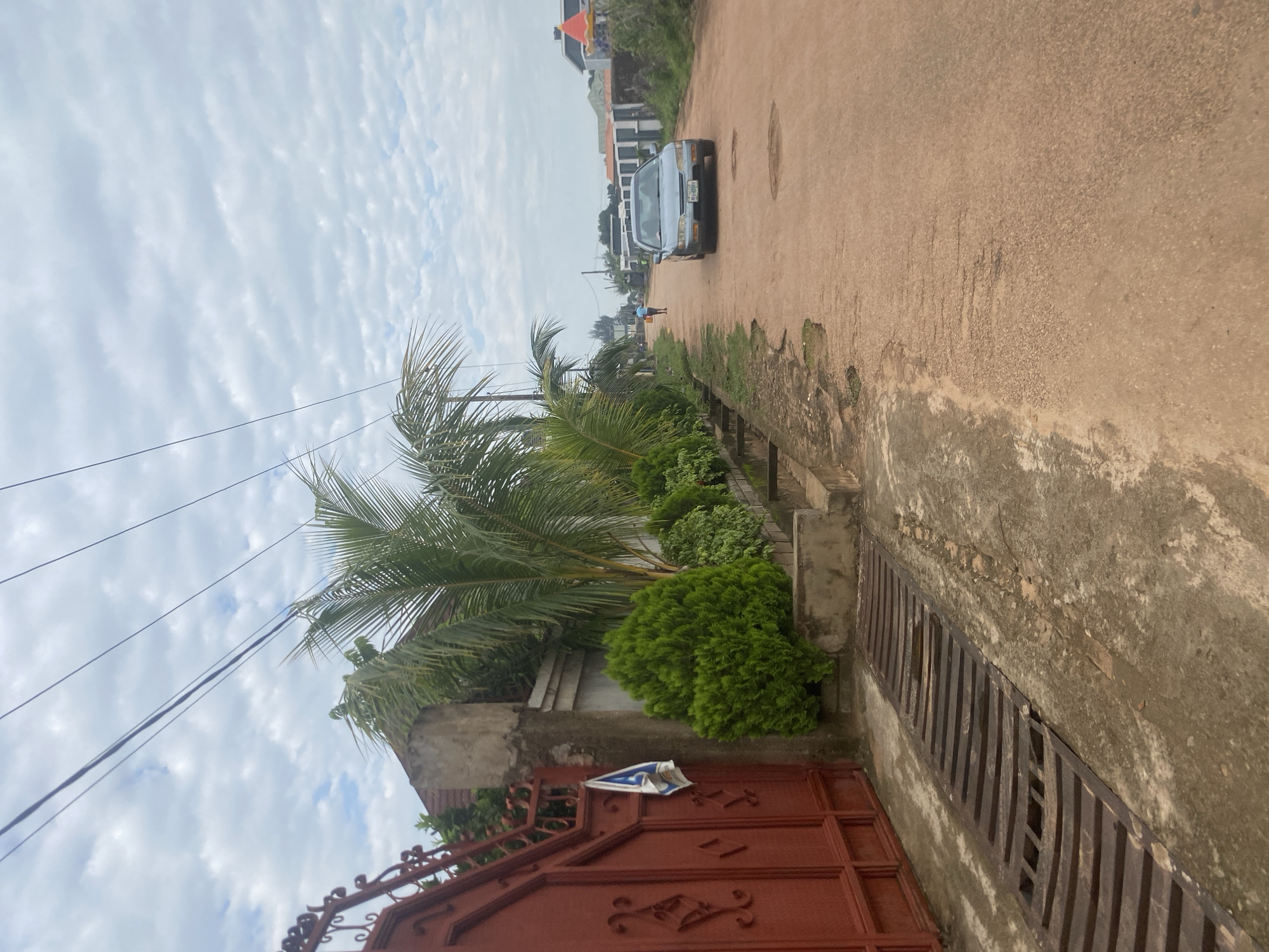 The image depicts a street scene in Barden Yana, Nigeria, under a cloudy sky. On the left side of the frame is a large, ornate red metal gate with decorative scrollwork. Adjacent to the gate, a concrete pillar supports the gate, and a white and blue cloth is hanging from it. In front of the gate and pillar is a wide metal grate covering a drain. Beyond the gate and pillar, a row of manicured green bushes and palm trees are visible, lined by a low concrete wall. The street itself is unpaved, with a reddish-brown dirt surface showing signs of wear and some water puddles. A light-colored sedan is driving away from the camera on the right side of the street. Further down the road, a person is walking away from the camera. In the background, several residential buildings with modern architecture are visible. Overhead, power lines crisscross the sky.