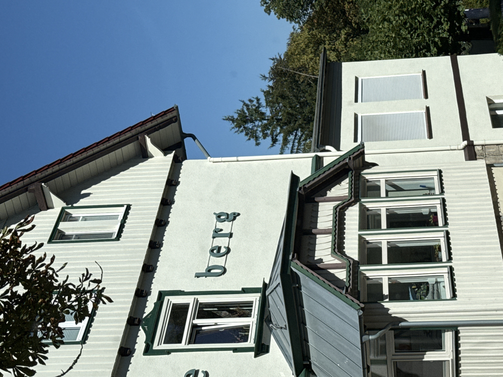 The image shows the exterior of a light green building, likely a house or guesthouse, with white trim and green accents. The building has multiple windows, some with white shutters. The name "berg" is visible in green lettering on the side of the building. The building is situated in a wooded area, with trees visible in the background. The sky is clear and blue. The architecture and the presence of the name "berg" suggest that this building is located in the town of Bad Sachsa, Germany, which is known for its natural beauty and is nestled in the Harz Mountains.
