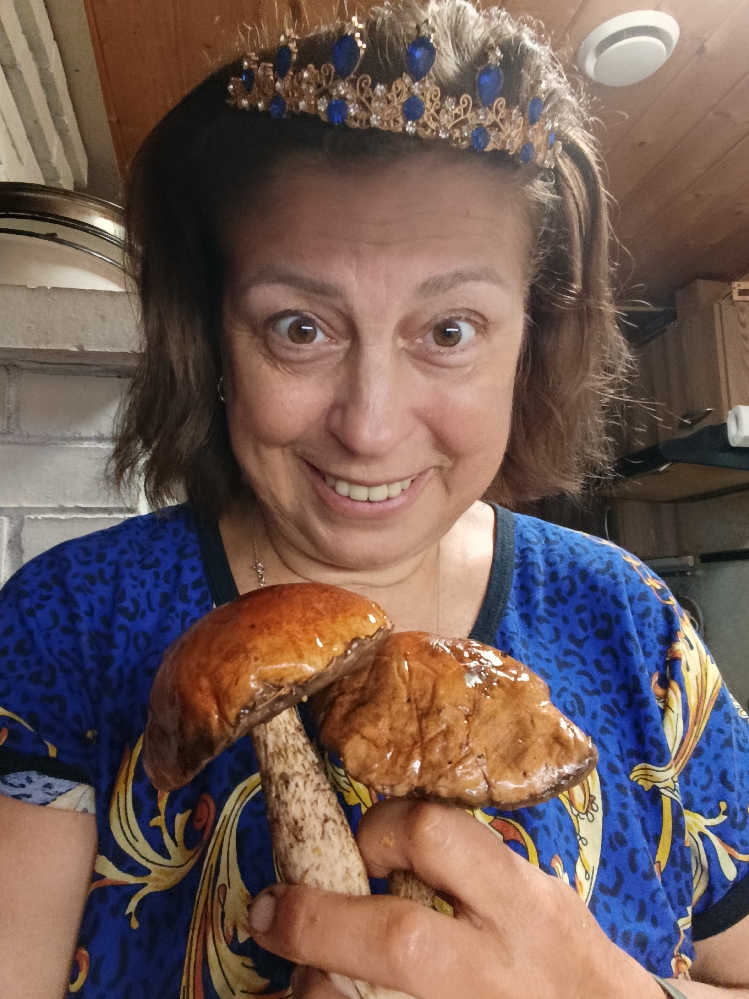 A woman with dark brown hair and a tiara adorned with blue gems smiles broadly as she holds two large mushrooms in her hands. She is wearing a blue shirt with a black leopard print and gold accents. The background appears to be a rustic kitchen with wooden paneling and exposed brick.