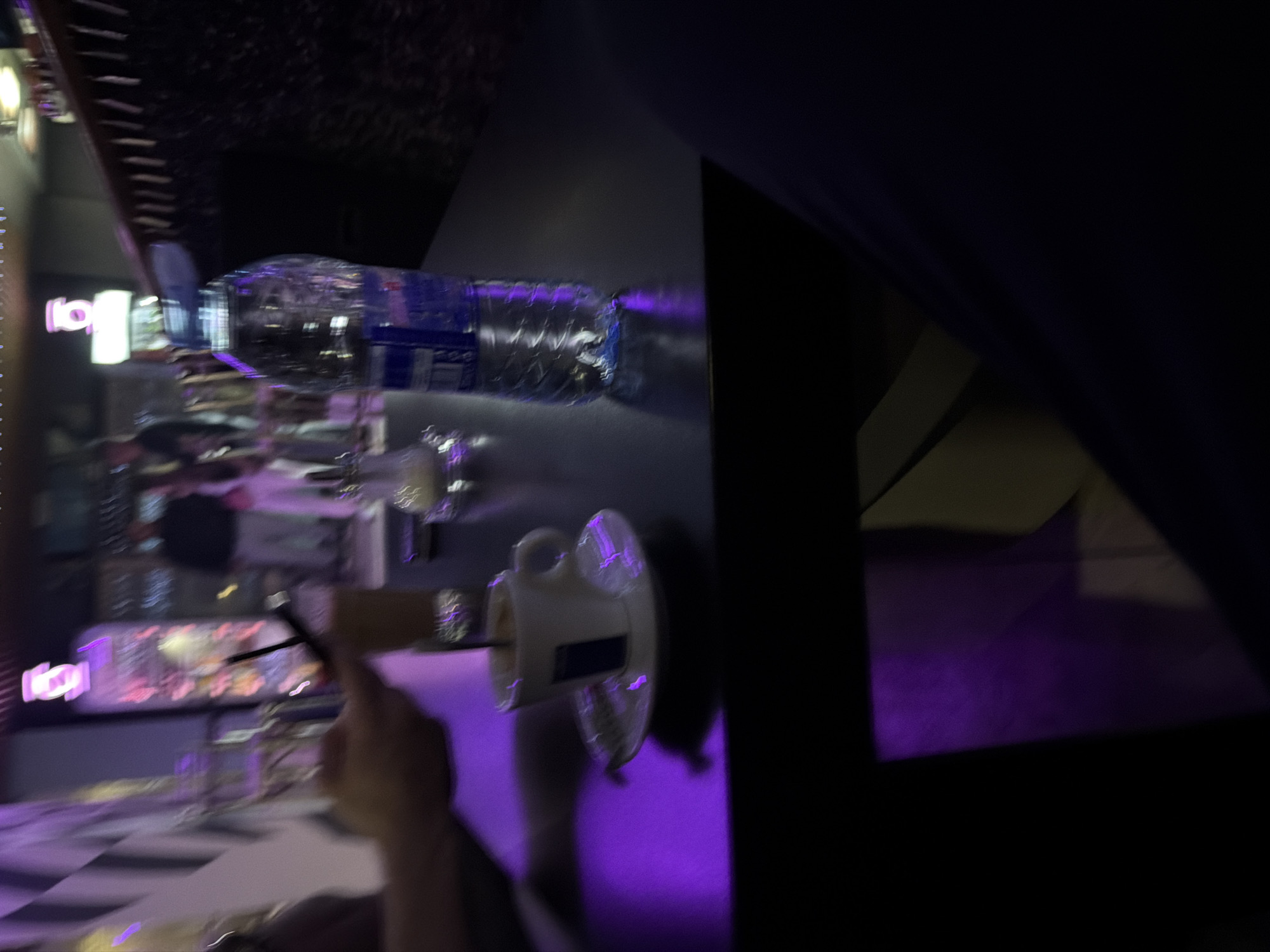 The image shows a blurred, low-lit indoor setting, possibly a restaurant or bar. In the foreground, a hand is visible, seemingly holding or manipulating a small, dark object like a pen or a thin tool. On the table, there's a mug or cup on a saucer and a plastic water bottle. The lighting appears to be violet or purple-toned. In the background, blurred neon signs and other tables/objects suggest an urban environment, however, the location is Unknown.