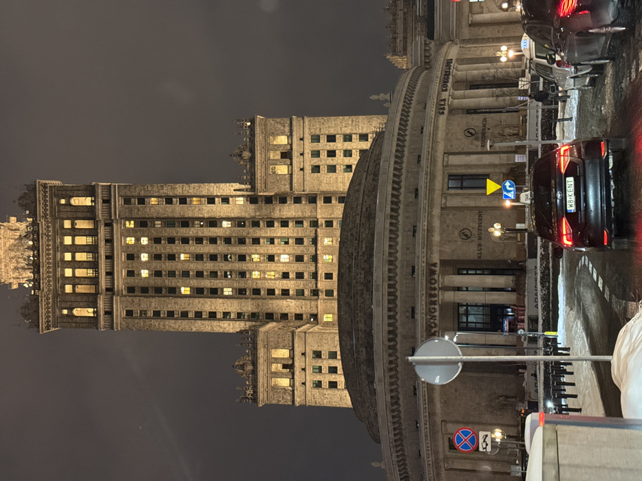 The image depicts a nighttime scene in Warsaw, Poland, showcasing the illuminated Palace of Culture and Science during winter. The colossal stone skyscraper, with numerous lighted windows and floodlit upper sections, dominates the background under an overcast dark sky. Its intricate architectural details, including smaller towers and ornate decorative elements, are visible.
In the mid-ground, a large, circular, multi-story stone building forms the base of the complex. This structure features prominent columns along its facade and signage that reads "KONGRESOWA" (Congress Hall) and "KLUB MUZYCZNY" (Music Club), with "SALA KONGRESOWA" also discernable.
The foreground presents a snow-dusted and slushy paved area, likely a road or parking lot. A black sedan, identified as a taxi by a roof sign and bearing the Polish license plate "WB KENT," is positioned centrally, facing away from the viewer, with its red taillights illuminated. A second vehicle, partially visible on the right, also appears to be a taxi.
Street furniture includes a red-bordered "No Stopping" sign (B-36), a yellow triangular "Yield" sign combined with a blue "Turn Right" arrow (D-1/D-4), and a blank circular sign on a pole. Several traditional-style streetlights cast yellow illumination. Bollards line the curb in front of the circular building. A single, indistinct person is visible walking on the sidewalk to the right. Snow covers the ground along the edges of the paved areas.