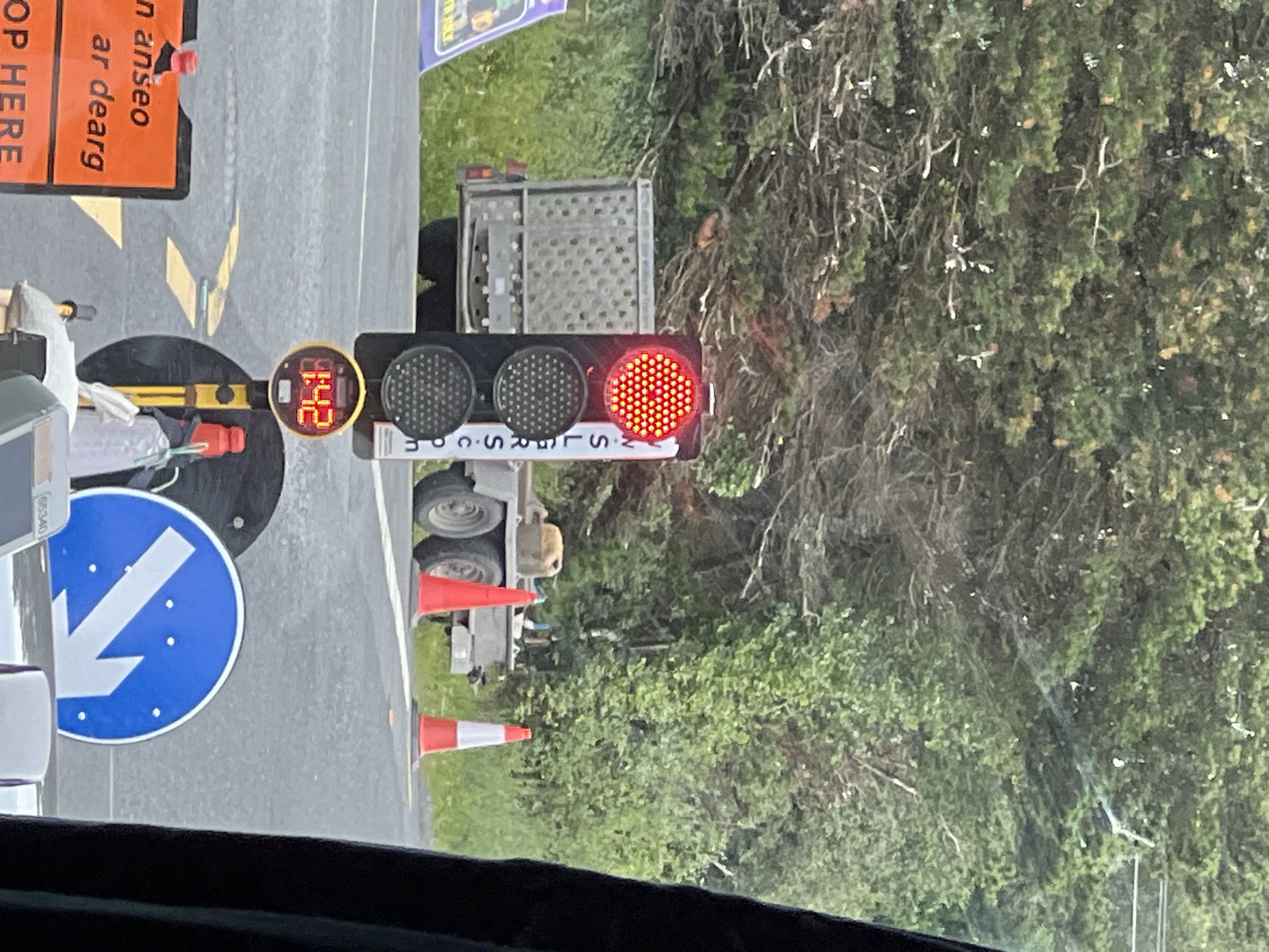 In Creeslough, Ireland, a temporary traffic light displays a red signal, indicating a stop. The traffic light is mounted on a pole and has three signal heads, with the top one illuminated red. Below the red light, two dark signal heads are visible. A digital timer is positioned below the traffic light, showing "1:42".
In the background, a small trailer with a cage-like structure is parked. Two traffic cones, one solid orange and the other orange and white striped, are placed on the road. Further back, a dense line of trees and foliage forms the backdrop.
In the foreground, an orange traffic sign with white lettering reads "n anseo ar dearg" and "OP HERE". To the right of this sign, a blue circular road sign with a white arrow pointing downwards and to the left is visible. Several people, mostly obscured by the vehicle the photo is taken from, are implied to be present, as suggested by the context of a traffic control situation. The overall scene depicts a temporary traffic management setup, likely due to road work or an event in Creeslough.