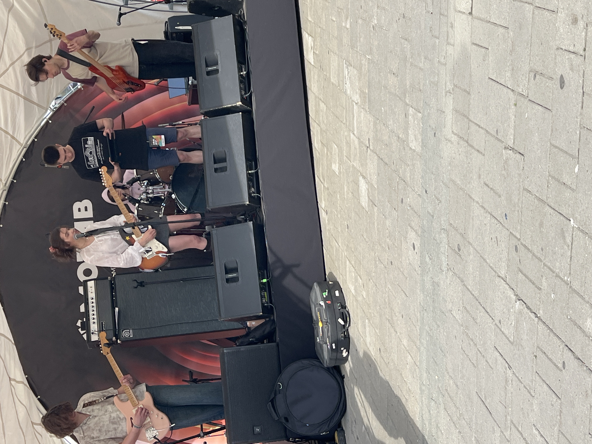 A band is performing on a stage in Yekaterinburg, Russia. The band consists of four musicians. From left to right, the first musician is a male playing a pink electric guitar and singing. The second musician is a female playing a white electric guitar and singing. The third musician is a male playing a black electric guitar and looking at a tablet. The fourth musician is a male playing a red bass guitar. The stage has black speakers and amplifiers. In front of the stage are two cases, one round and one rectangular, possibly for musical equipment. The ground is paved with light gray square and rectangular tiles, and there are shadows on the ground. The background shows a black banner with red and white graphics and text. A white tent is visible above the stage.