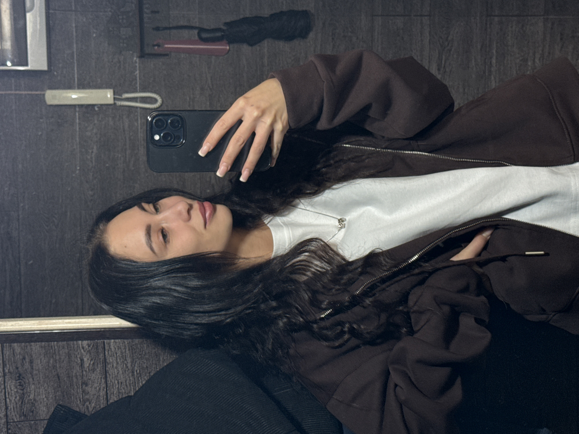 An indoor mirror selfie captures a young woman from the chest up, reflected in a large mirror. She has long, dark, wavy hair and is seen in a slight profile, looking towards her reflection. She is wearing a dark brown zippered hoodie over a plain white t-shirt, with a thin silver necklace visible against the white fabric. Her lips have a subtle light-colored lipstick, and her facial expression is neutral.
Her right hand holds a dark-colored smartphone, likely an iPhone given the distinct triple camera lens array visible on its back. Her fingers, which have neatly manicured nails with a French tip style, are spread across the phone.
The background, visible as a reflection in the mirror, consists of a dark, textured surface, possibly wood paneling or large dark tiles. Reflected items include a light-colored object resembling an intercom receiver with a coiled cord, a black umbrella hanging, and a reddish-brown horizontal object, perhaps a shelf or rail, behind the umbrella. The setting appears to be an interior space, possibly a hallway or room within a building in Chaikovsky, Russia. There is no visible text in the image.