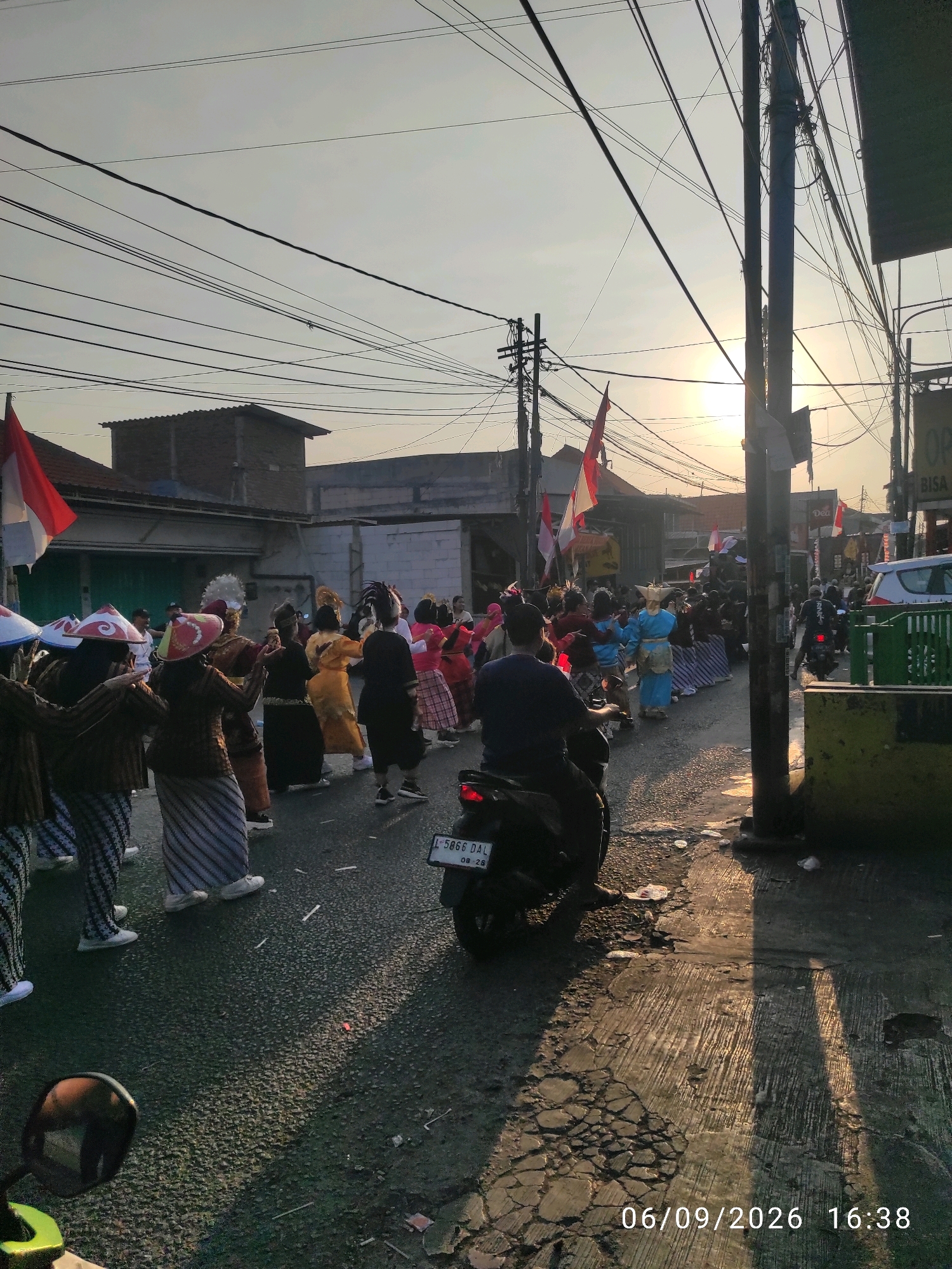 A group of people in traditional clothing are participating in a parade on a street in Wage, Indonesia. The sun is setting, casting long shadows across the road. The people are wearing colorful outfits and some have elaborate headdresses. They are walking in a procession, and some are holding flags. There are also a few motorcycles in the scene, with one person riding a motorcycle in the foreground. The street is lined with buildings, and there are power lines overhead. The date and time stamp on the image reads "06/09/2026 16:38".