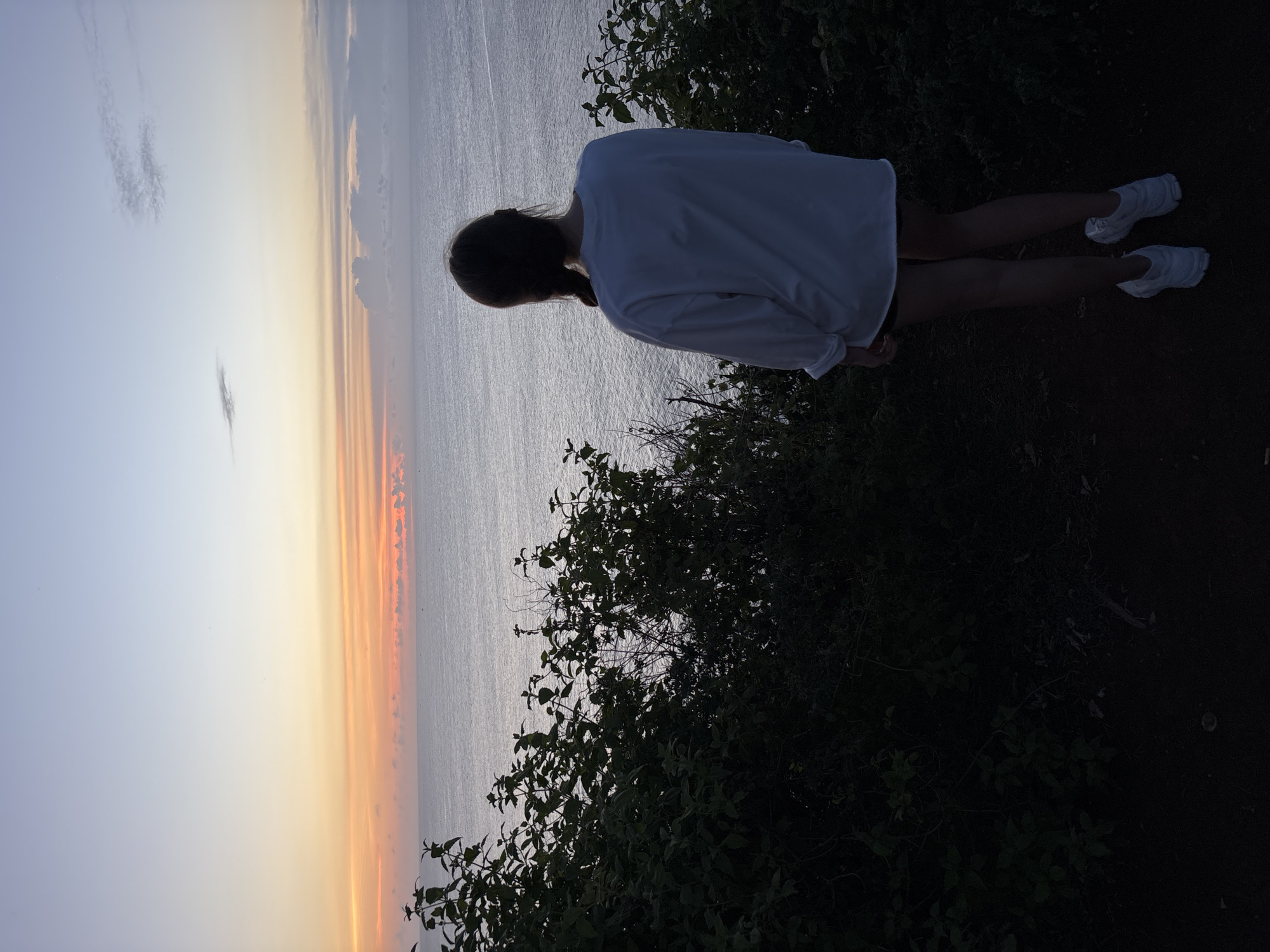 A woman with braided hair stands at the edge of a cliff, overlooking the ocean. She is wearing a white, long-sleeved shirt and white sneakers. The foreground features dark foliage, suggesting a natural environment. Beyond the woman and the foliage, the ocean extends towards a vividly colored sunset, with hues of orange, yellow, and blue. The location is Pecatu, Indonesia, based on the likely environment and the sunset coloration.