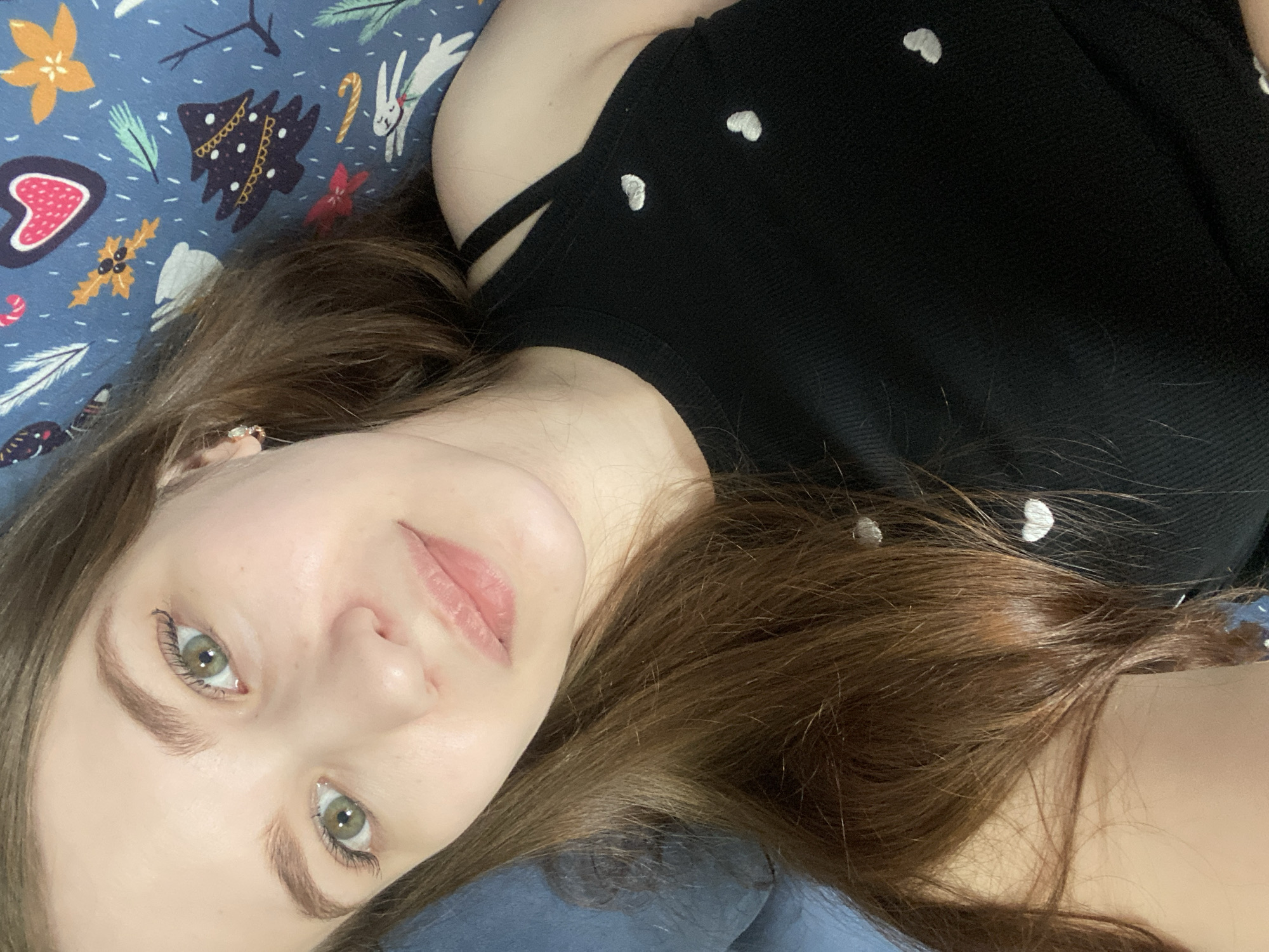 The image displays a close-up, high-angle portrait of a young adult female, located indoors in the city of Chaikovsky, Russia. She is positioned lying down, with her head resting on a plain dark blue surface, likely a pillow, situated to the left. Her long, brown hair is visible, extending around her head and partially over her right shoulder and chest. The subject has a fair complexion, green eyes with visible eyelashes, and light brown eyebrows. A small red mark is discernible near the inner corner of her right eye. Her lips are naturally pink and slightly parted. A small, light-colored stud earring is present in her right ear. She is attired in a black, ribbed tank top with thin shoulder straps. This garment features several small, white, heart-shaped embellishments on its front. To the right of the subject and behind her upper body, a blue fabric with a repetitive pattern of various holiday and winter-themed motifs, including Christmas trees, hearts, candy canes, and rabbits, is visible. The female subject is looking directly into the camera with a neutral to slightly serene facial expression.