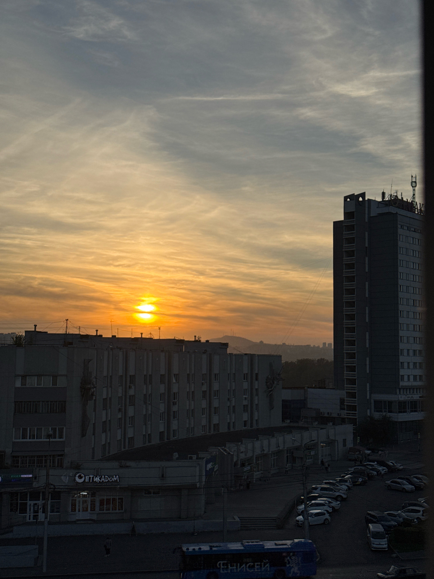 The image depicts an urban scene at dusk or dawn, with the sun low on the horizon, casting a warm orange and yellow glow across the sky and clouds. Several buildings dominate the foreground and midground. A tall, dark, rectangular building with many windows stands prominently on the right. To its left, a lower, multi-story building with a lighter facade features multiple windows and architectural details. In the foreground, a street level view shows a row of parked cars, a blue bus with Cyrillic lettering, and several pedestrians. One building has a sign with Cyrillic text that appears to read "ОПТИКАDOM". The location is identified as Krasnoyarsk, Russia.