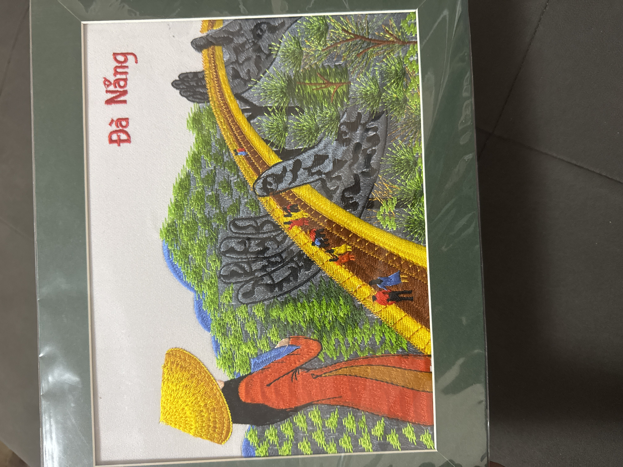 The image shows a framed embroidery of a scene. The scene depicts a mountain pass in Da Nang, Vietnam, based on the text "Đà Nẵng" in the upper left. A yellow road winds between gray rocky mountain sides covered with green vegetation. Small figures are embroidered along the road. In the foreground, the embroidered depiction includes a figure with a conical Asian-style hat and reddish-orange clothing. The image is a close up of the embroidery in a dark green frame, and is angled slightly toward the viewer. The background shows a dark gray surface. There is no location in the city of Plovdiv, Bulgaria.