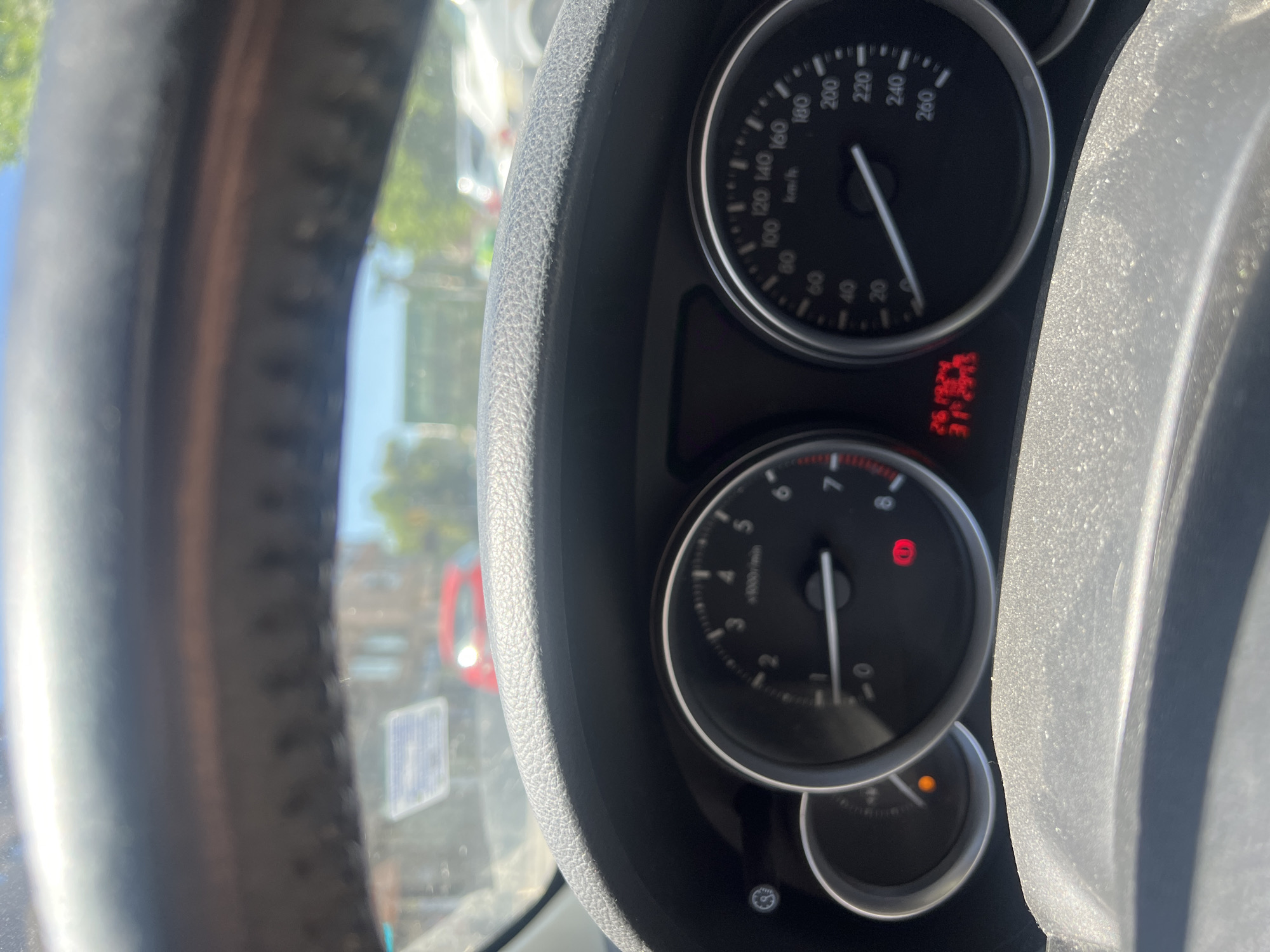 The image shows the dashboard of a car. The speedometer reads approximately 110 km/h, and the tachometer needle is near the 0 mark. The odometer shows 261324. Warning lights are illuminated, including a red light with an exclamation point inside a circle and an orange light indicating a possible issue with the engine or exhaust system. A portion of the steering wheel is visible on the left. The scene is inside a car, presumably in Burgas, Bulgaria, though the city's location cannot be definitively determined from the dashboard alone. Visible through the windshield are blurry shapes which possibly indicate an urban environment with streetlights and the back of a red car.