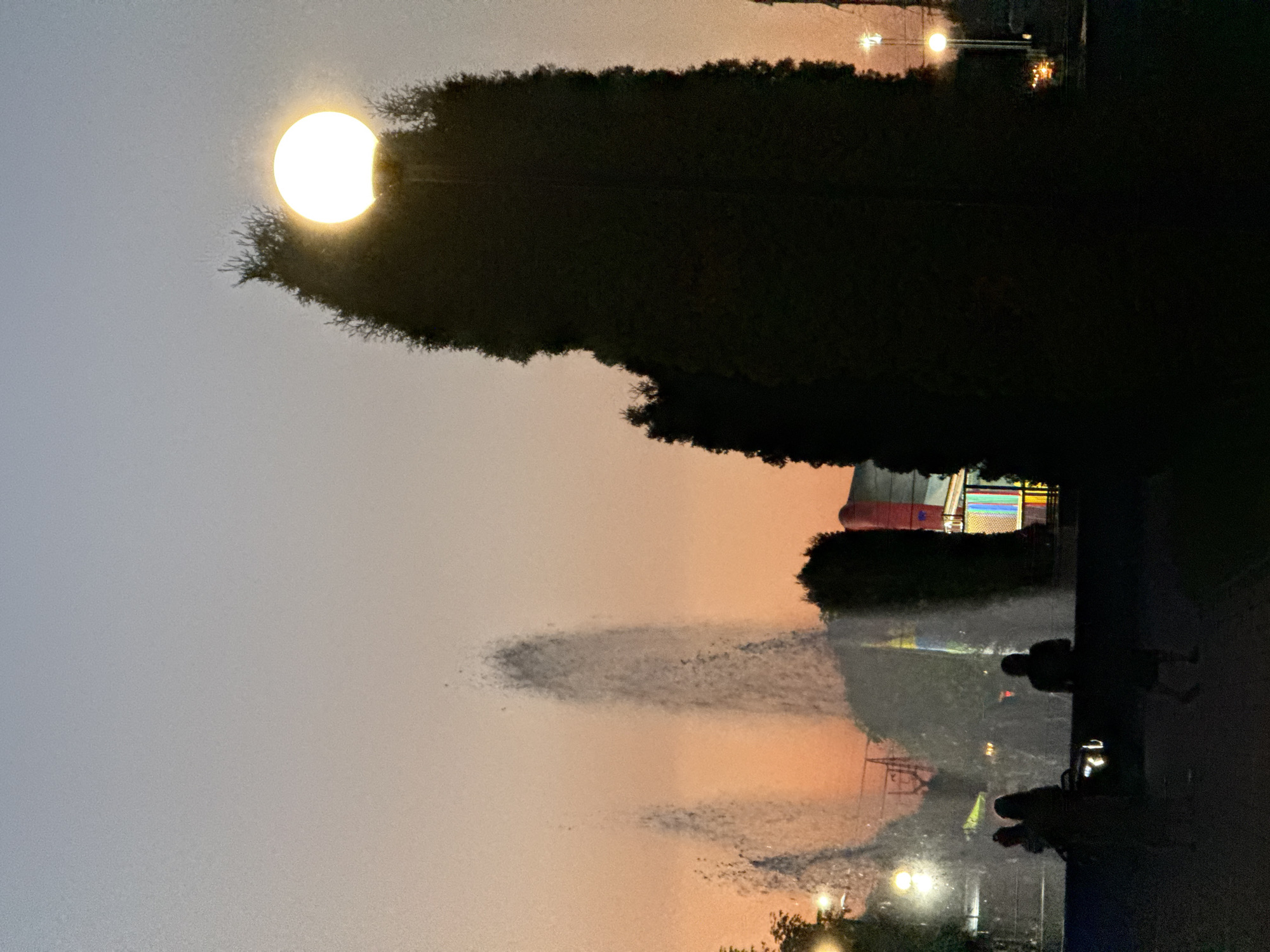 The media file depicts an outdoor scene at dusk in Endirey, Russia. The sun, a bright, illuminated orb, is partially obscured by the dark silhouette of a dense tree line in the foreground. The sky transitions from a pale gray on the left to a warm orange hue on the right, indicating the sunset.
In the background, beyond the trees, a brightly lit structure resembling a fairground attraction with red and white striped awnings is visible. Several figures, silhouetted against the fading light, can be seen walking or standing. The ground appears wet, with reflections suggesting recent rain or a water feature, such as a fountain, which is creating misty plumes of water. Streetlights are also visible, casting a warm glow. The overall mood is serene and peaceful, capturing the transition from day to night. No visible text is present in the image.