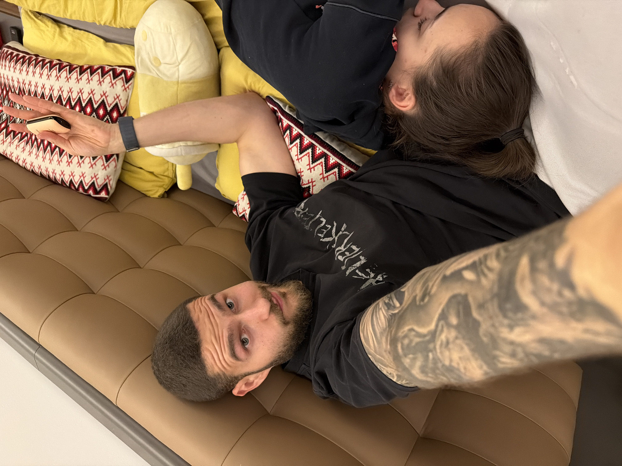 This indoor photograph captures two individuals relaxing on a brown, tufted couch or bench in Sofia, Bulgaria. The man, positioned in the foreground and closer to the camera, lies on his back with his head slightly tilted, looking directly at the viewer with a neutral, slightly inquisitive expression. He has short dark hair, a beard, and wears a black t-shirt with stylized white text, possibly "KRELEPIR" or similar, across the chest. His left arm, prominently displayed in the shot, is covered in a detailed grayscale tattoo.
Behind the man, a woman is visible, lying on her side with her face mostly obscured, appearing to be resting or asleep. Her dark hair is pulled back in a low ponytail secured with a black elastic. Her right hand, extended across decorative pillows, holds a small, light-colored electronic device, possibly a vape or a compact phone. She also wears a dark grey smartwatch on her left wrist.
The setting includes several pillows: two solid yellow ones and one with a distinctive red, black, and white geometric pattern. An elongated, light-colored plush toy or pillow is also visible beside the woman. The overall scene suggests a casual and relaxed atmosphere, possibly in a lounge area or a comfortable public space.