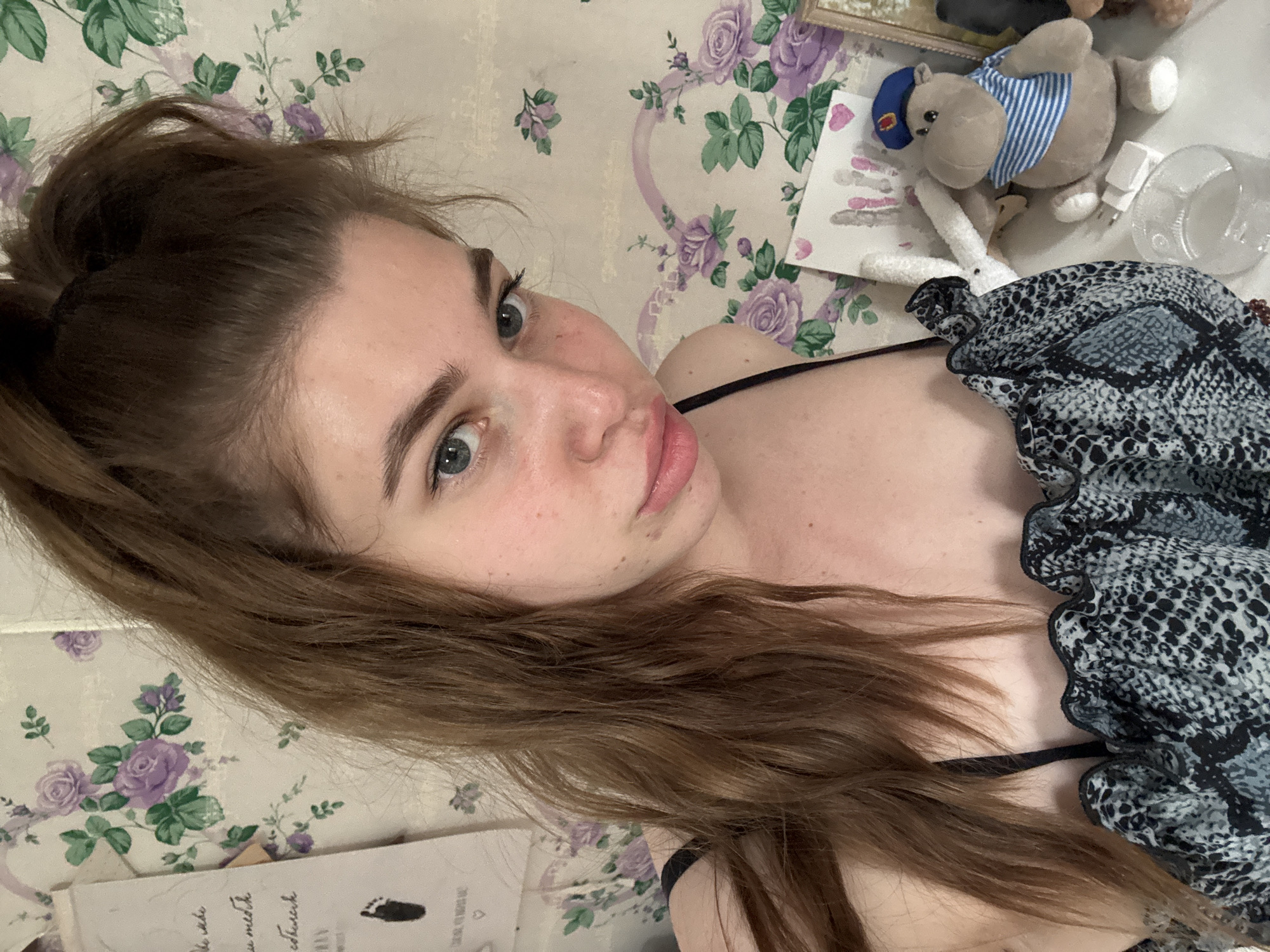 A young woman with fair skin and brown hair partially tied up poses for a selfie indoors. Her eyes are light-colored, and she has freckles on her face. She is wearing a black top with a patterned fabric draped over her chest and shoulder. The background consists of a wall covered with a floral pattern featuring purple roses and green leaves. On a surface next to her, there are several items, including a stuffed hippopotamus wearing a sailor hat and a striped shirt, a stuffed white rabbit, a framed photo, a piece of paper with a drawing, and a clear plastic container. The location is not identifiable as Мещёры, Russia, based on the image alone.