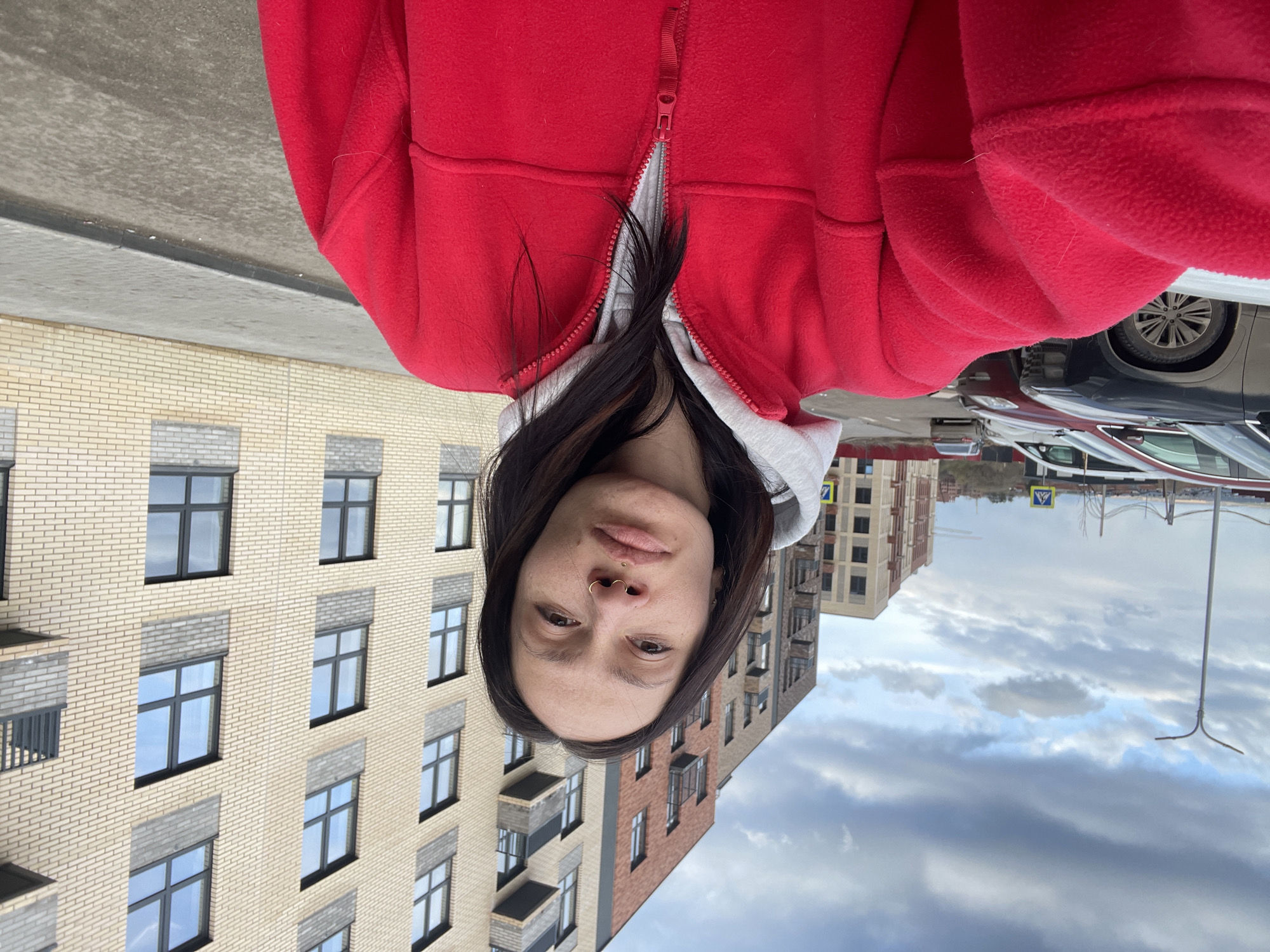 The image displays a young woman, likely in her 20s or 30s, looking directly at the viewer. She has long, dark hair and is wearing a red fleece jacket, which is partially unzipped, revealing a lighter-colored (possibly grey or white) hoodie underneath. A gold septum piercing is visible on her nose.
The setting is an urban environment, likely a residential area in Izhevsk, Russia. In the background, there are multi-story apartment buildings; one on the left is made of light beige brick, and another on the right features a mix of beige and darker brown/red brickwork. These buildings have numerous windows with dark frames and some balconies. A street or sidewalk is visible, with several parked cars. The sky is overcast with clouds, suggesting a cloudy daytime. A street pole with a pedestrian crossing sign (a white triangle with a black walking figure on a blue background) is faintly discernible in the distance.
The woman's expression is neutral and calm. The original image is rotated 90 degrees clockwise, presenting the woman and the immediate foreground upside down relative to the background buildings and sky. No specific readable text is visible, only the symbolic pedestrian crossing sign.
