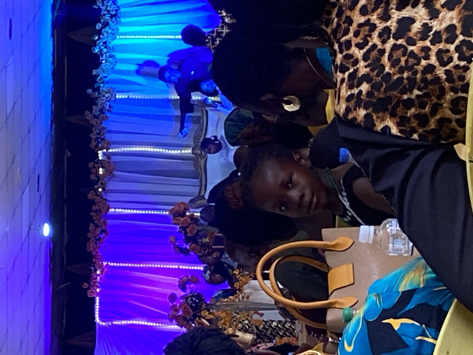 A sideways, indoor shot shows a young girl looking at the camera, amidst a crowd of people seated in what appears to be an event hall in Gura Top, Nigeria. The girl is positioned slightly to the left of the center, and she's wearing a black and white patterned top. Behind her is an adult woman in a leopard print top, partially visible.
There are various objects scattered in front of the girl, including a tan-colored handbag, and a water bottle.
In the background, stage decorations in blue and purple hues, with what appears to be flowers along the edges, can be seen. There's also a faint image of people sitting behind the girl.