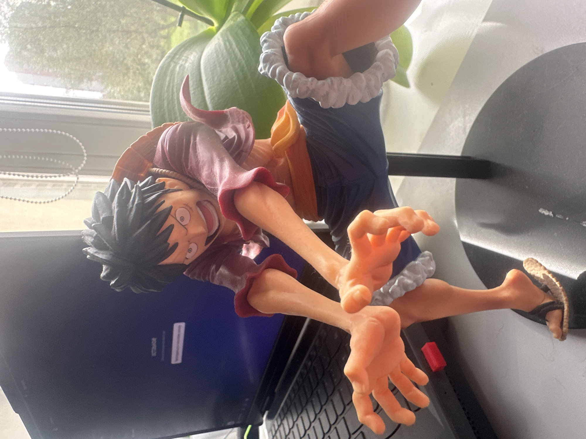 The image shows a close-up of a Monkey D. Luffy action figure, a character from the anime and manga series One Piece. The figure is seated on a black stand, which is placed on a desk next to a laptop. The figure is wearing his iconic red shirt with rolled-up sleeves, an orange sash, and blue shorts with a white trim. His arms are outstretched and his hands are open, as if he is about to grab something. His legs are bent and one foot is clad in a straw sandal. In the background, a window reveals trees and buildings, suggesting an urban setting. A string of white beads hangs in front of the window. The scene appears to be indoors, possibly in a home or office. There is no indication of the specific location in Maladzyechna, Belarus, beyond the implied presence of an urban environment.