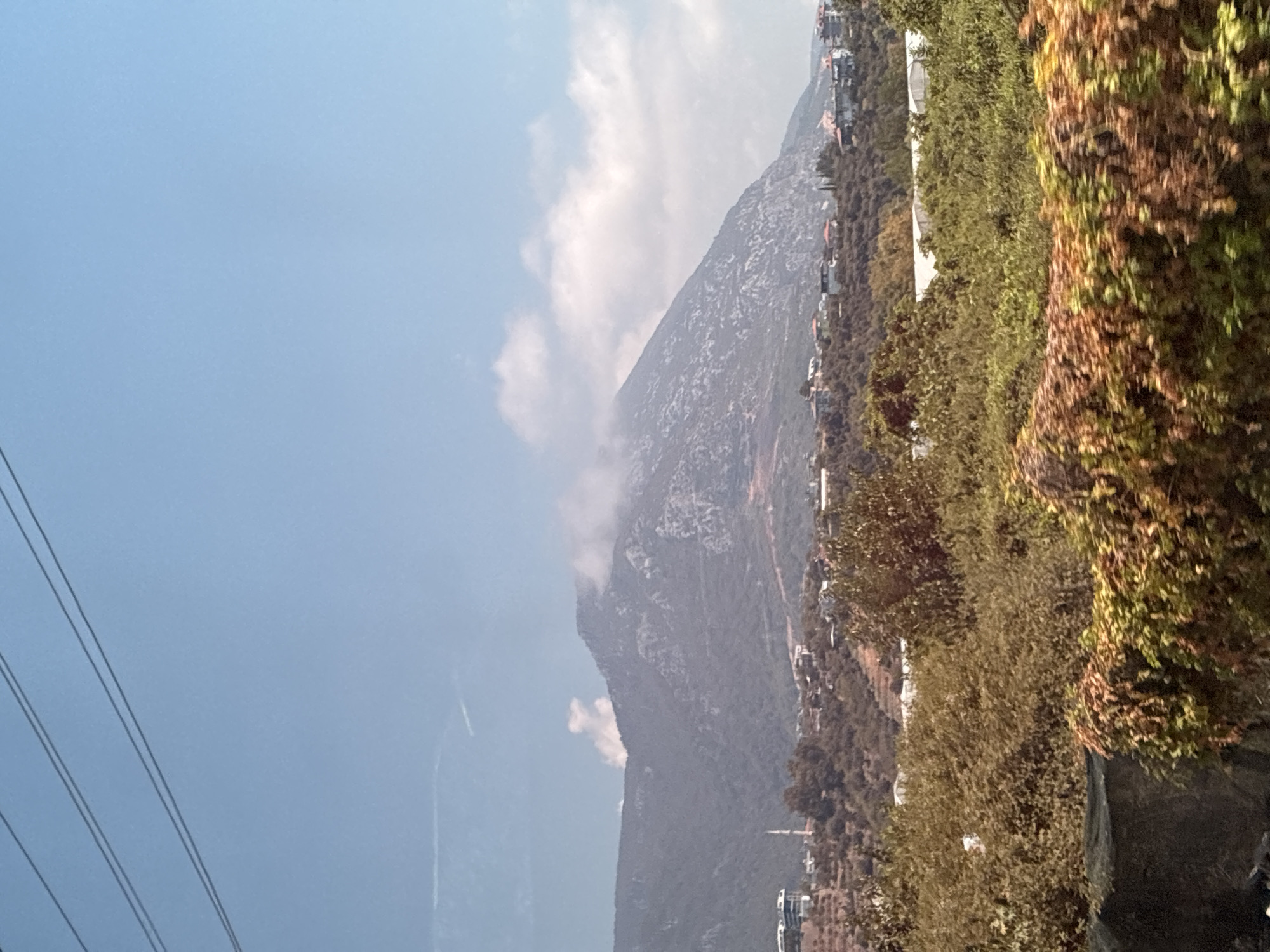 The image depicts a mountainous landscape with a town nestled at its base, under a clear blue sky with scattered clouds. In the foreground, dense, leafy vegetation with a mix of green and brown hues dominates, with some climbing vines visible. A dark, textured surface, possibly a wall or embankment, runs along the very bottom edge of the frame.
In the middle ground, a town is spread across the lower slopes of the mountain. Buildings with red-tiled roofs are visible amongst trees and other vegetation. A slender minaret, characteristic of a mosque, stands out amongst the buildings on the left side of the town. The mountain itself rises sharply in the background, with its rocky surface partially obscured by wisps of cloud near its peak. Three power lines are strung across the upper portion of the sky.
This scene is consistent with the topography and urban development found in Alanya, Turkey, particularly in areas that border the Taurus Mountains. The presence of a minaret further reinforces a likely location in Turkey. No people are visible in the image. The environment appears to be a daytime scene, likely during warmer months due to the lush vegetation.