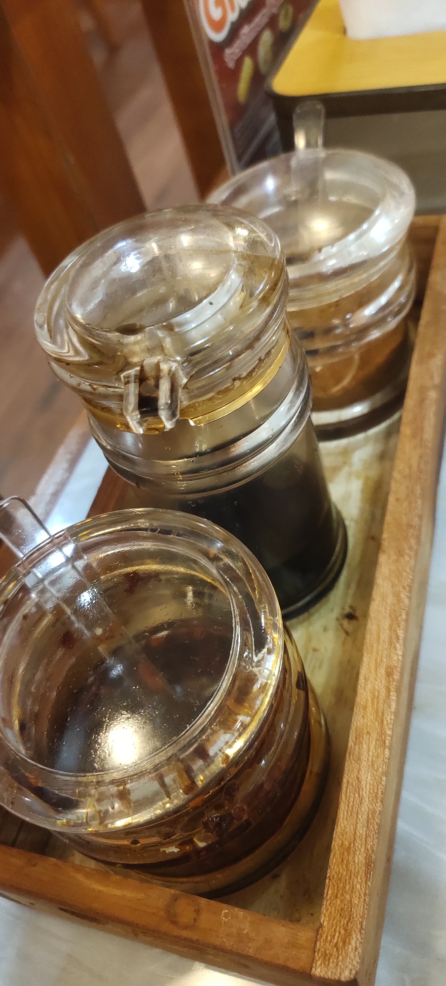 This is a close-up, top-down, outdoor shot of three glass condiment shakers in a wooden tray. The shakers are filled with dark brown liquid, possibly soy sauce or chili oil. The lighting suggests it's daytime, and the setting appears to be a restaurant or food stall in Kudus, Indonesia. The tray is resting on a light-colored surface, likely a table. The focus is on the condiment shakers and their contents, with a blurry background suggesting other elements of the dining environment.