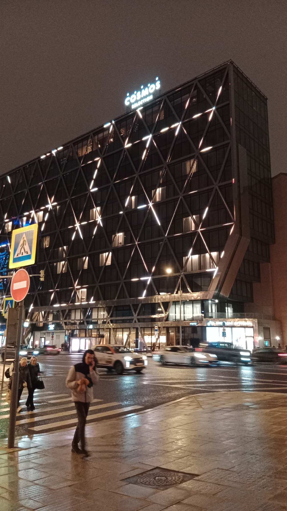 The image shows a street scene at night in Moscow, Russia, featuring the Cosmos Selection hotel. The hotel is a modern building with a facade characterized by a pattern of intersecting triangles and illuminated lines. The hotel's name, "Cosmos Selection," is prominently displayed in illuminated letters on its roof.
In the foreground, pedestrians are crossing a street with crosswalk markings. A man in a light-colored jacket and dark pants is walking and talking on a mobile phone. Two other people are visible to his left. Traffic, including several cars, is moving across the street. Streetlights illuminate the scene, and reflections are visible on the wet pavement. Road signs, including a crosswalk sign and a "no entry" sign, are present.