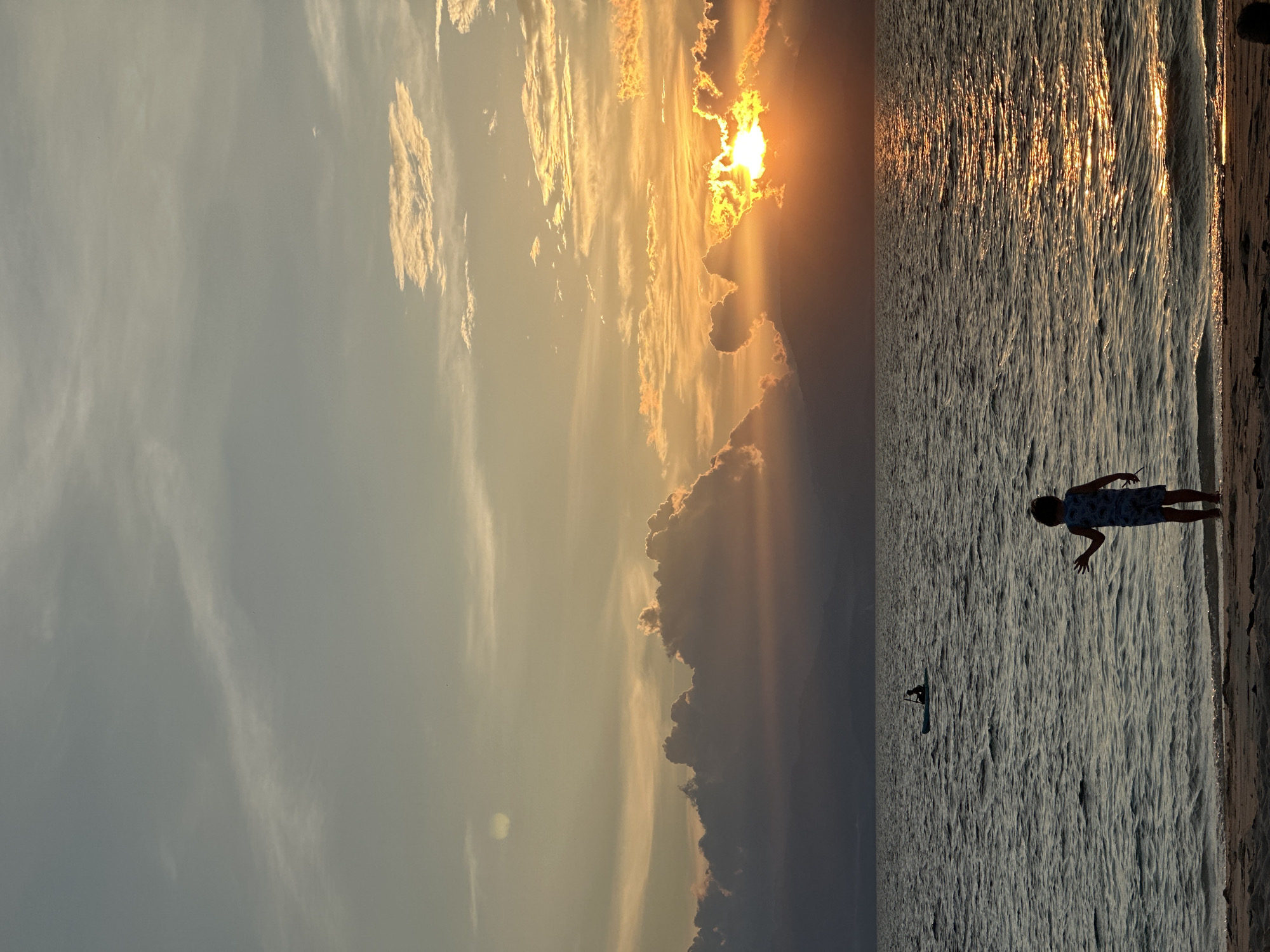 The image depicts a serene sunset scene on a beach in Saavedra, Philippines. The sky is filled with a gradient of colors, from pale yellow and orange near the horizon to a soft blue higher up, with scattered clouds. The sun, partially obscured by clouds and mountains in the distance, casts a brilliant golden glow on the rippling water.
In the foreground, a child stands at the water's edge on a sandy beach, facing the ocean and raising one hand as if waving. The child is silhouetted against the sunset. Further out in the ocean, a lone figure can be seen paddling a small boat, possibly a kayak or paddleboard. The water appears calm, with gentle waves lapping at the shore. The overall atmosphere is peaceful and tranquil, capturing a moment of natural beauty and quietude.