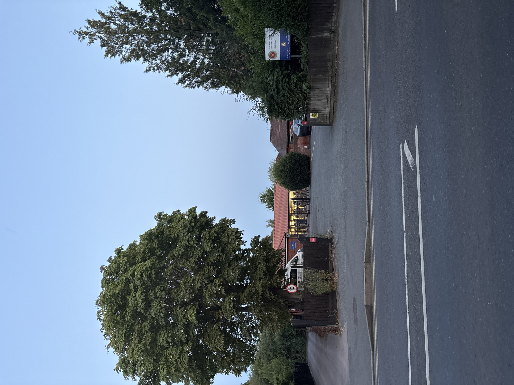 The image depicts the entrance to the East Anglian Reserve Forces and Cadets Association in Chelmsford, United Kingdom. In the foreground, a grey asphalt road with white lane markings is visible. Beyond the road, a paved area with a wooden fence separates the road from the facility. A large tree with green foliage dominates the left side of the image, casting a shadow. A white van with "10 CDI SQUADRON" written on its side is parked behind the fence, near a speed limit sign indicating "10". Several people in military uniforms are standing in formation near a building with a red roof on the right side of the image. A sign with the crest of the East Anglian Reserve Forces and Cadets Association and information about its services is mounted on a wooden fence. Two cars are parked within the facility's grounds. The sky is a clear, pale blue, suggesting it is either early morning or late evening. The overall scene suggests a military or cadet training facility.