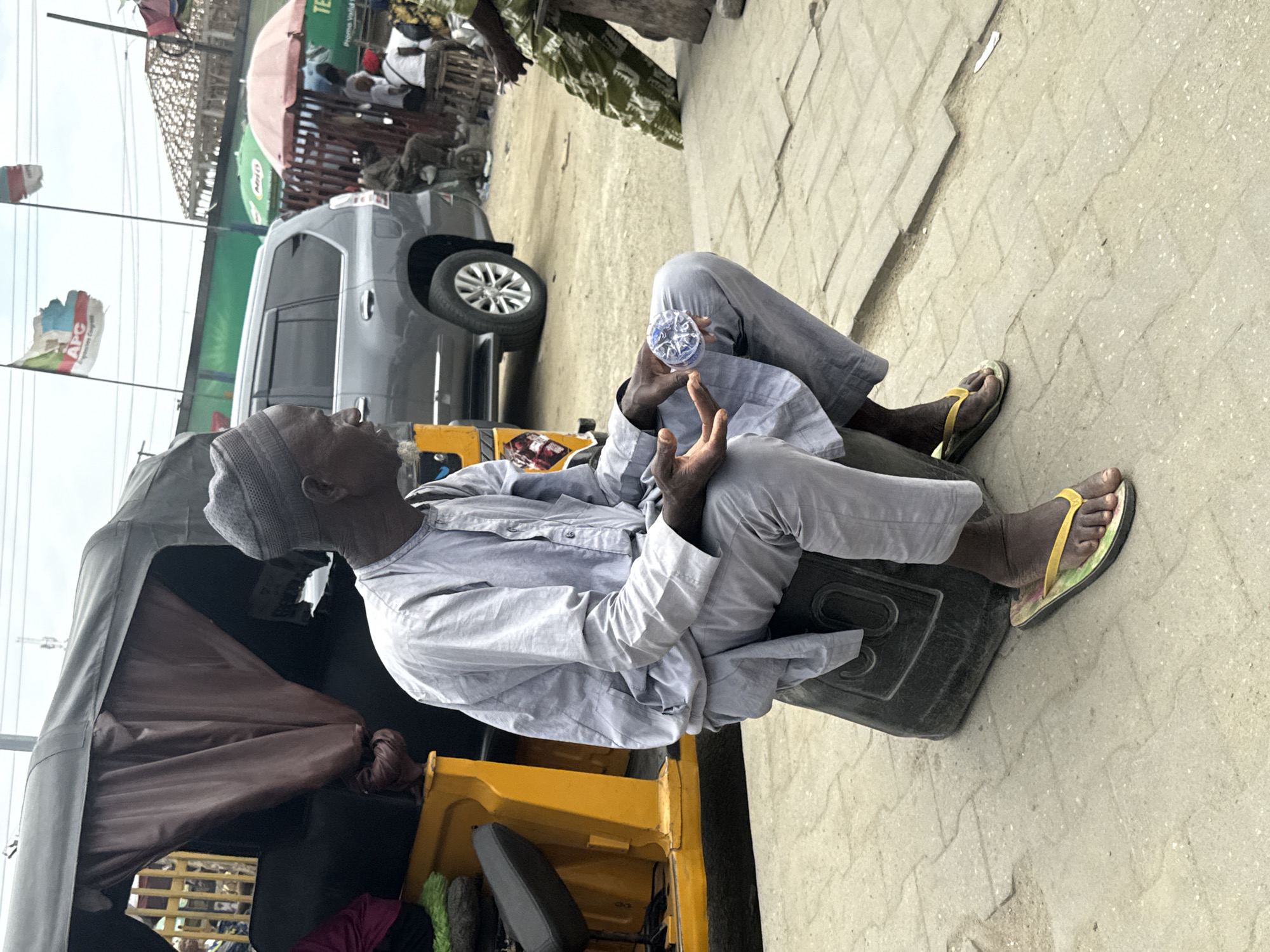 A man wearing a grey cap and grey traditional attire sits on a black rectangular object on a paved walkway in Lekki, Nigeria. He is holding a small, round, clear plastic object with a blue and white circular pattern inside it. To his left, a portion of a yellow and grey auto-rickshaw is visible. In the background, a silver SUV is parked, and further behind, there are various stalls with umbrellas and people milling about. Two flags, one with the "APC" logo, are visible in the distance. The scene appears to be an outdoor market or street area.