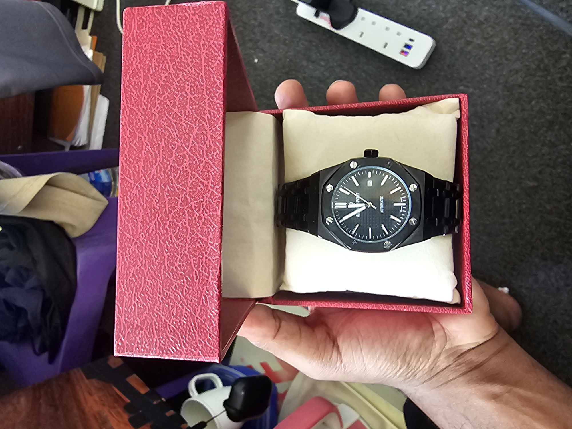 A person's hand is holding open a red textured box. Inside the box, nestled on a white cushion, is a black wrist watch with a black metal band. The watch face is black with silver hour markers and hands. The word "AUTOMATIC" is visible at the bottom of the watch face. To the right of the box, a white remote control with several buttons is visible. In the background, there are various items including a white mug with a black handle, a purple object, and some papers. The scene appears to be indoors. There is no visible indication of the specific location within the city of Dar es Salaam, Tanzania.