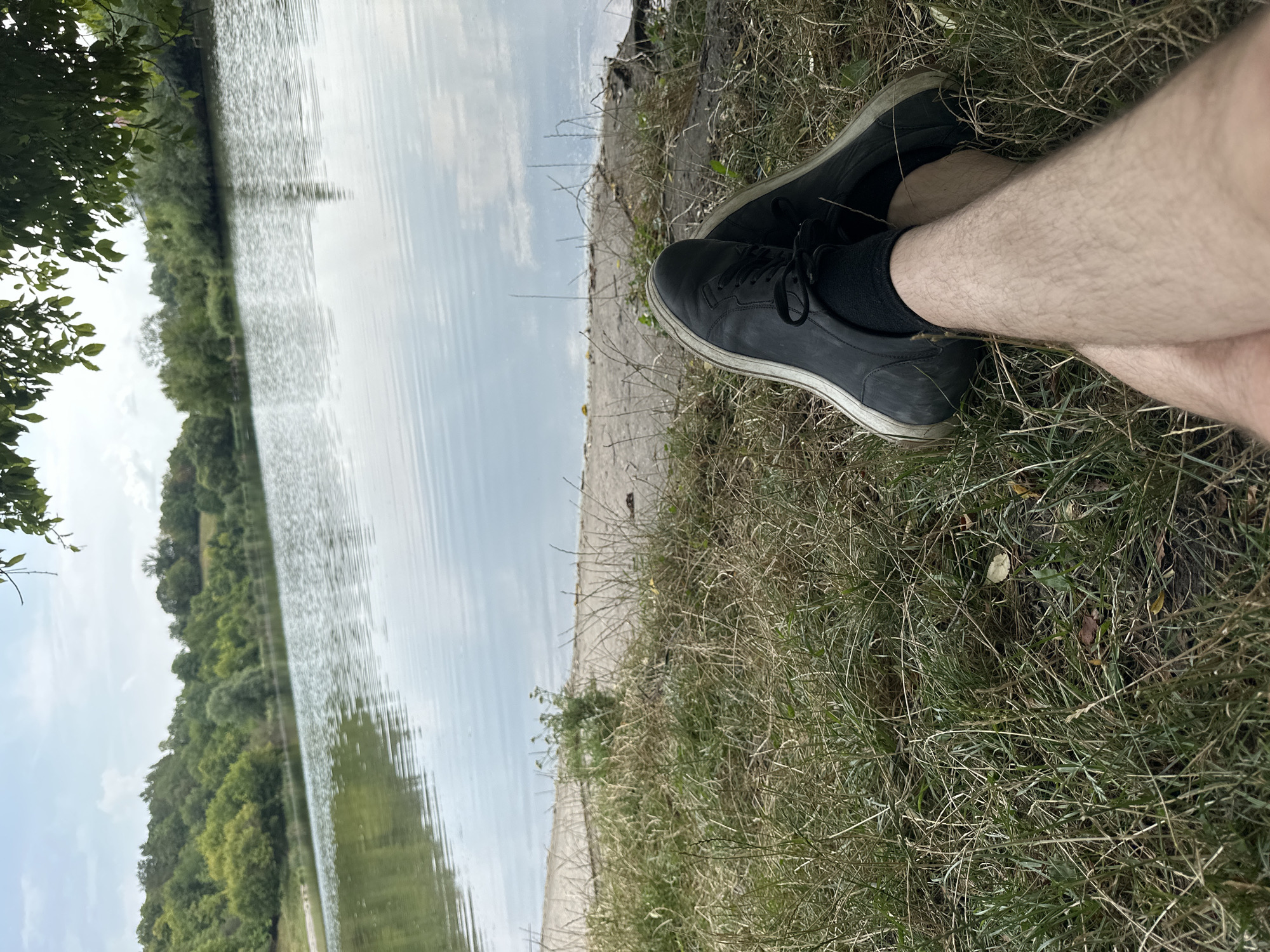 A pair of black sneakers and legs with hairy skin are resting in the foreground on a patch of dry grass near the edge of a calm body of water. The water reflects the sky, which has scattered clouds, and a green, treed hillside across the lake. A concrete path runs along the edge of the water behind the grass. The scene appears to be an outdoor recreational area.